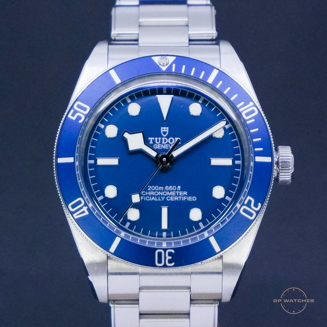 A close-up of a blue Tudor Geneve wristwatch with a stainless steel band, featuring a blue dial, white markers, and a unidirectional bezel with minute markings, and the text '200m:660ft', 'CHRONOMETER', and 'OFFICIALLY CERTIFIED' on the face.