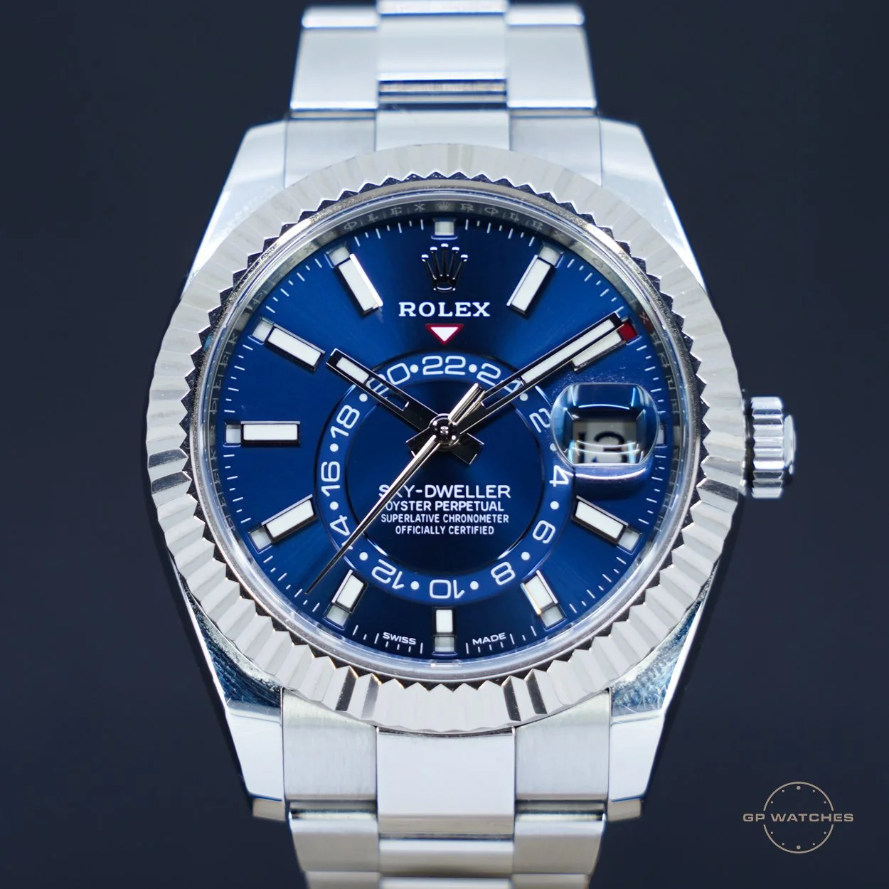 A close-up of a stainless steel Rolex Sky-Dweller watch with a blue dial, white markers, and a date window at 3 o'clock, against a dark background.
