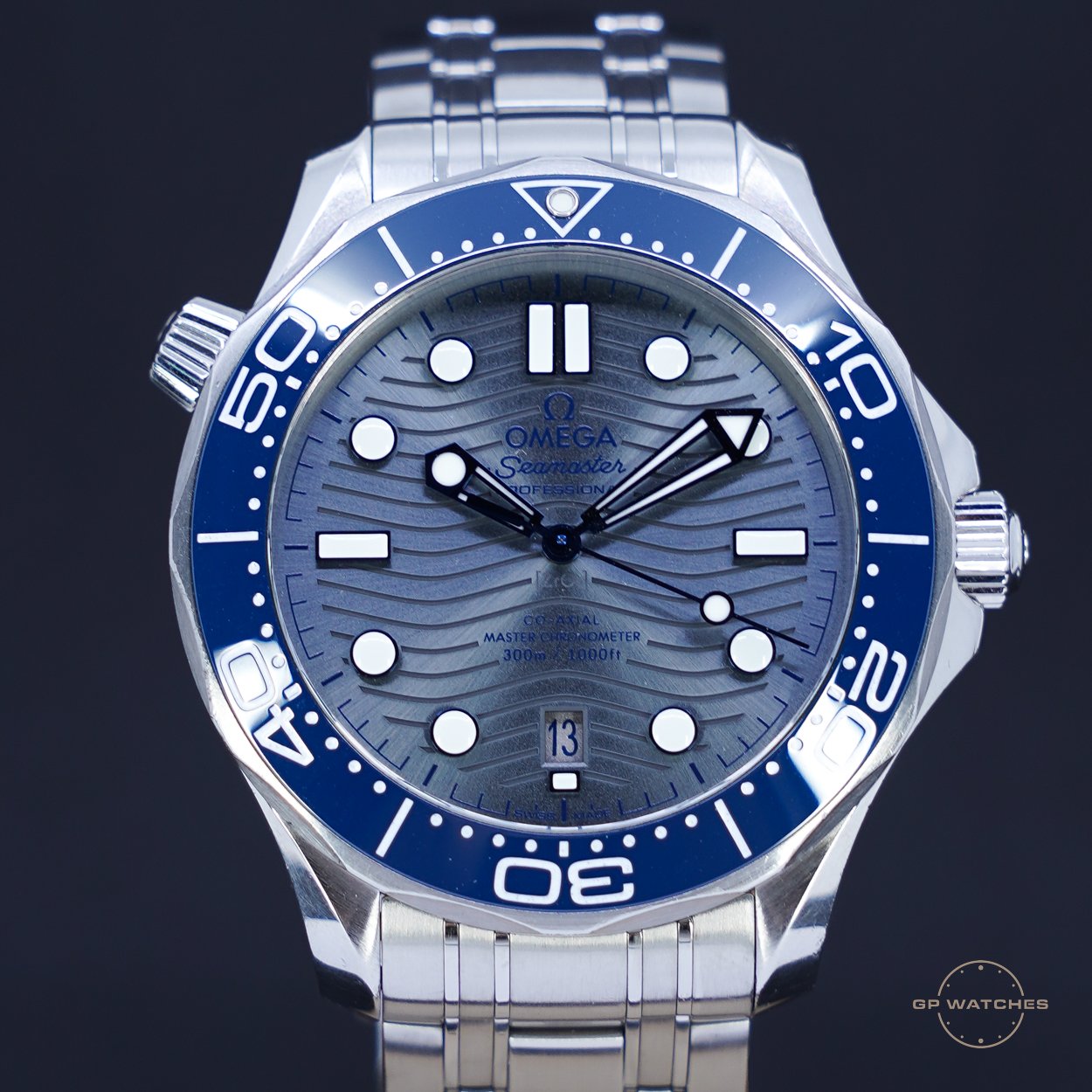 A close-up of a silver and blue Omega Seamaster wristwatch with a metal band, displaying the time as 8:20 and 14 seconds, with a date window showing the 13th on a dark background.