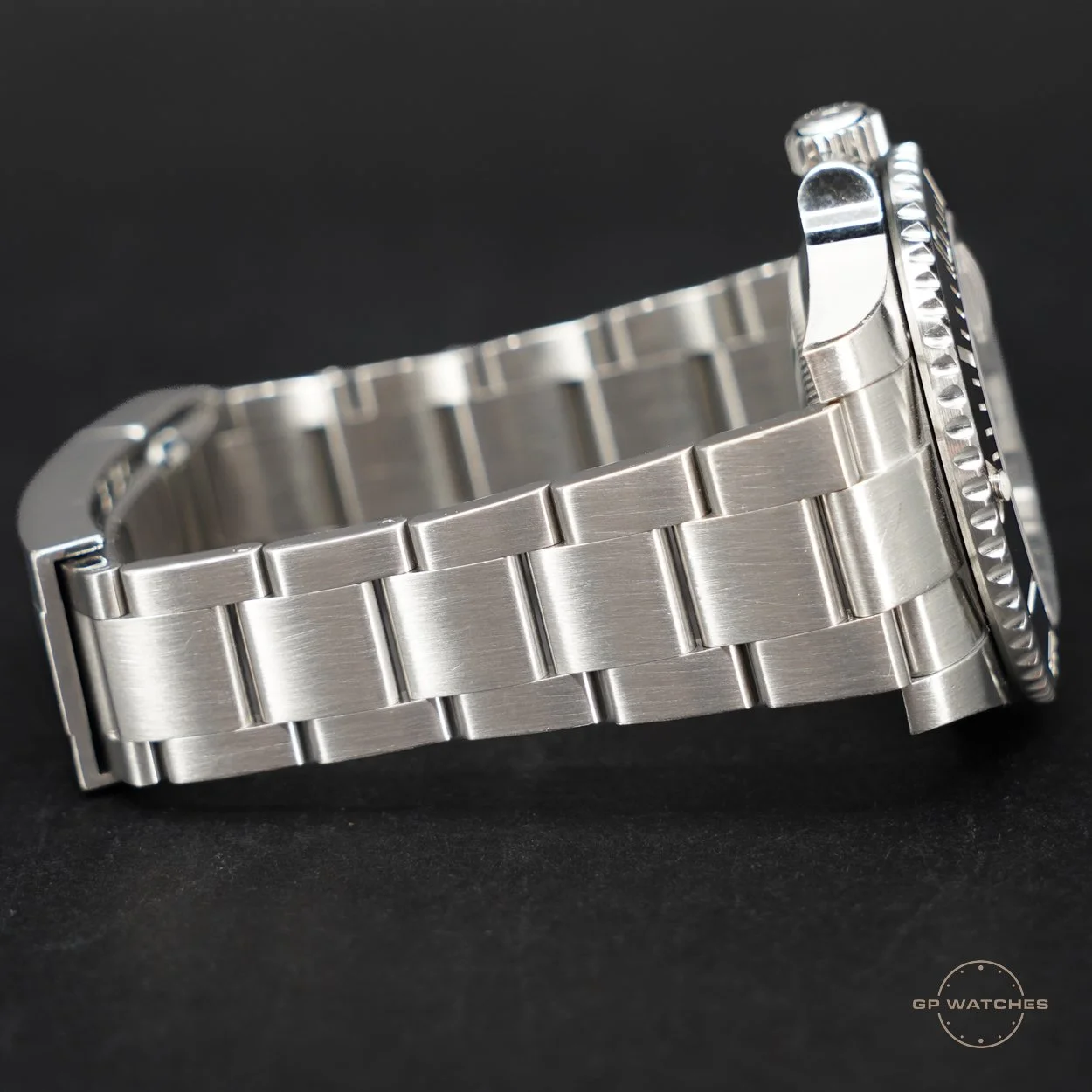 Close-up of a stainless steel watch bracelet with a brushed finish, resting on a black surface, with a partial view of the watch case and bezel.