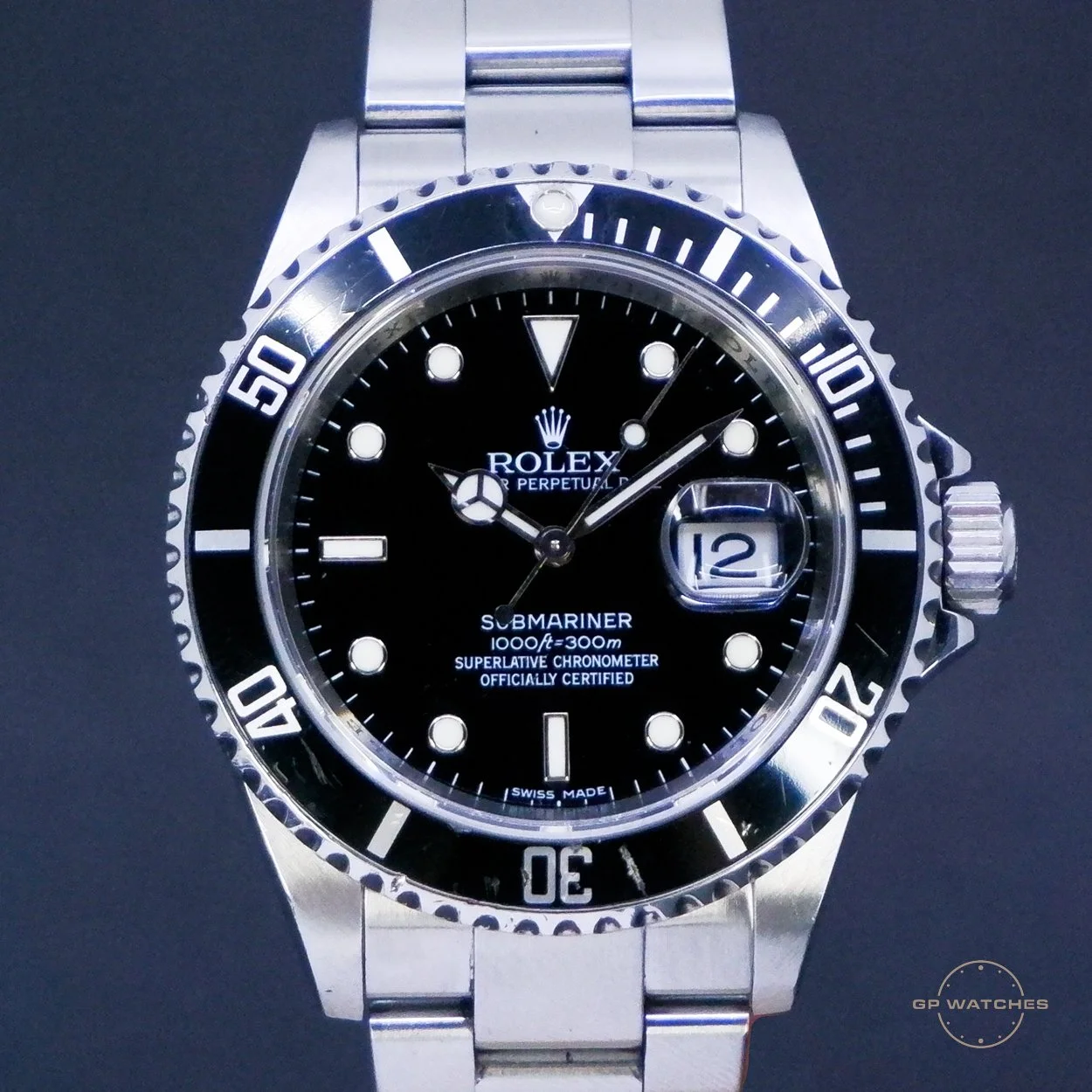 Close-up of a black dial Rolex Submariner wristwatch with a stainless steel bracelet. The watch features a unidirectional bezel, luminous hands and markers, a date window at 3 o'clock with a magnifying lens, and subtext indicating it is a superlative chronometer. The background is black.