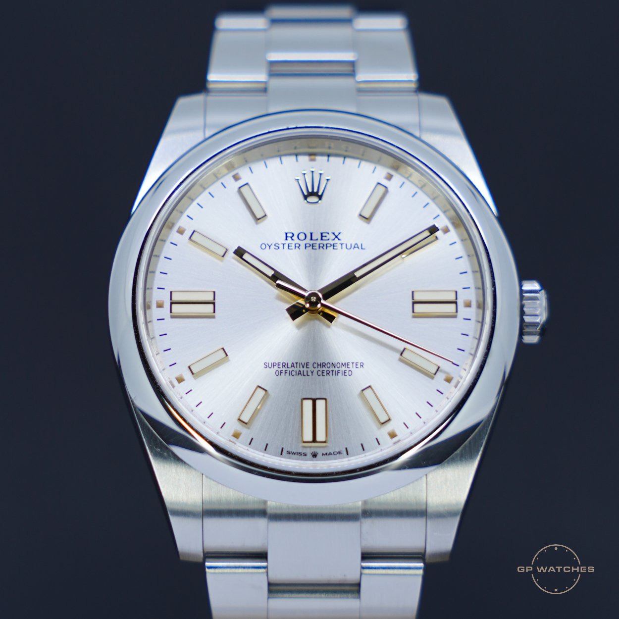 A close-up of a stainless steel Rolex Oyster Perpetual wristwatch with a silver dial, gold hour markers, and black hour and minute hands, against a black background.