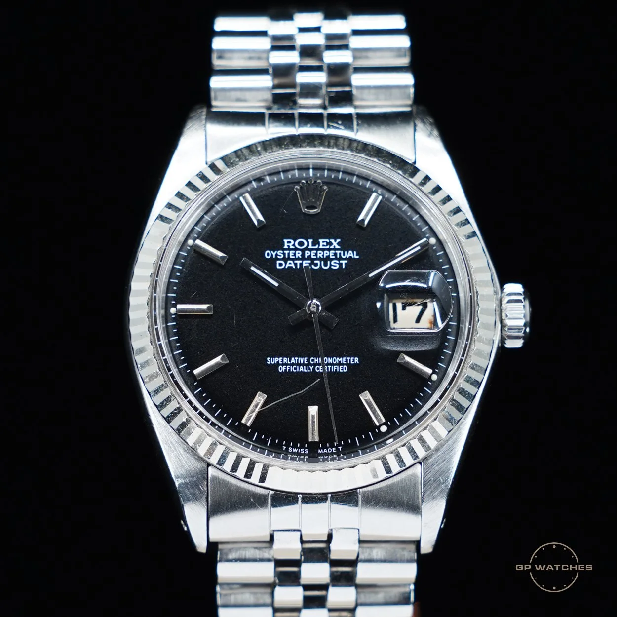 Close-up of a silver Rolex Oyster Perpetual Datejust wristwatch with a black dial, date display, and jubilee bracelet, against a black background.