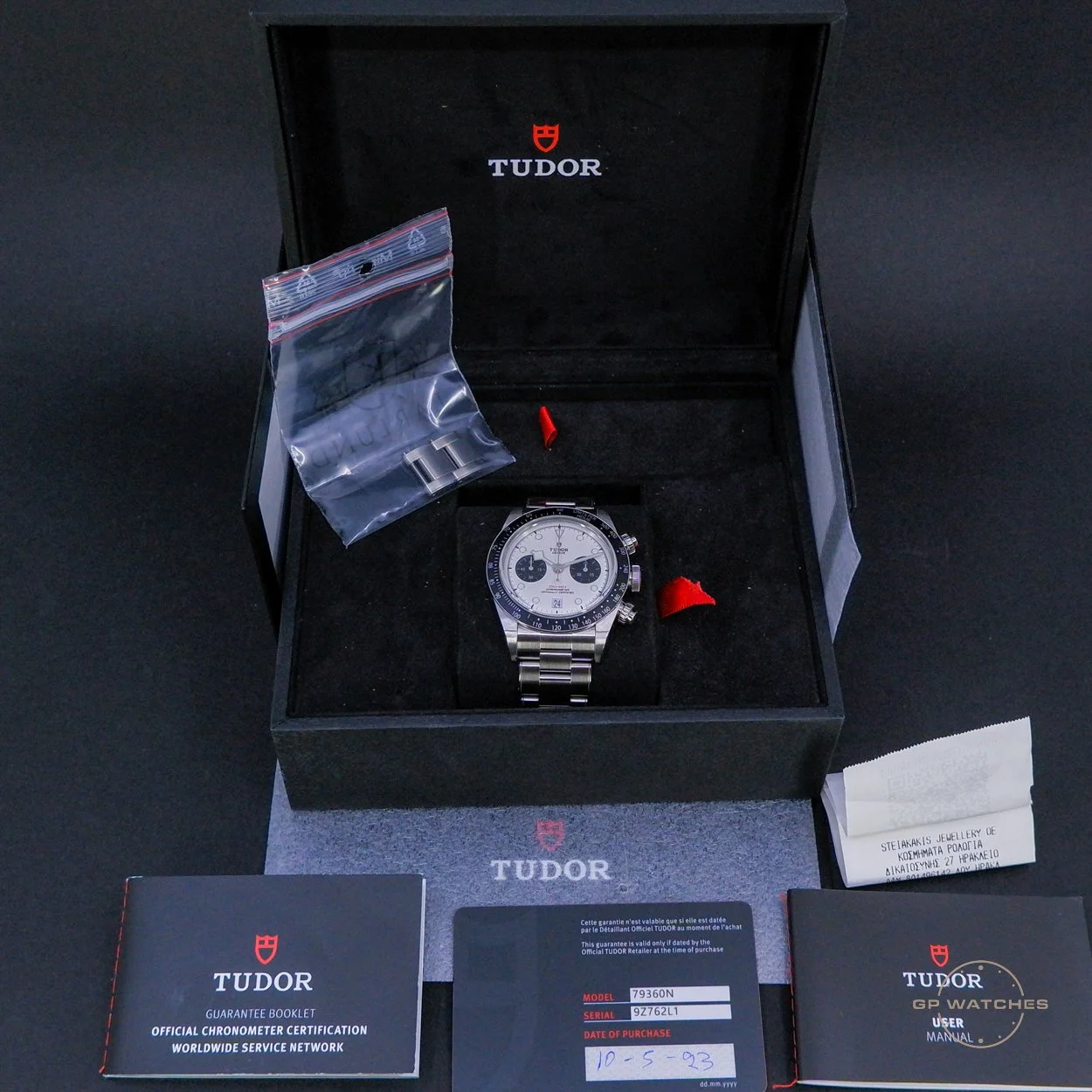 A Tudor wristwatch in its black box, with a stainless steel band and white dial featuring two subdials. The box contains an additional red plastic part and a small plastic bag with metal links. Surrounding the box are warranty and certification booklets, a serial number card, and a small white paper note, all on a black surface.