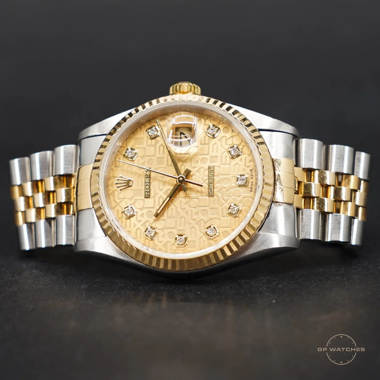 A luxury Rolex wristwatch with a two-tone metal band, a gold bezel, and a cream-colored patterned dial with diamond hour markers and a date window at 3 o'clock, set against a black background.