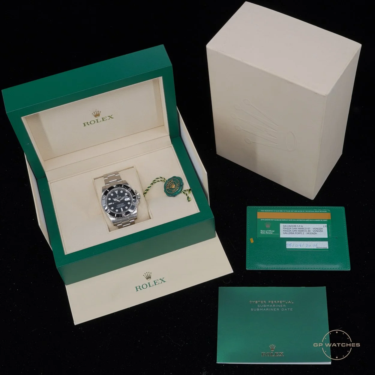 A Rolex Submariner watch inside its green and beige box, with accompanying certificates and a white outer box with embossed Rolex crown logo, all placed on a black surface.