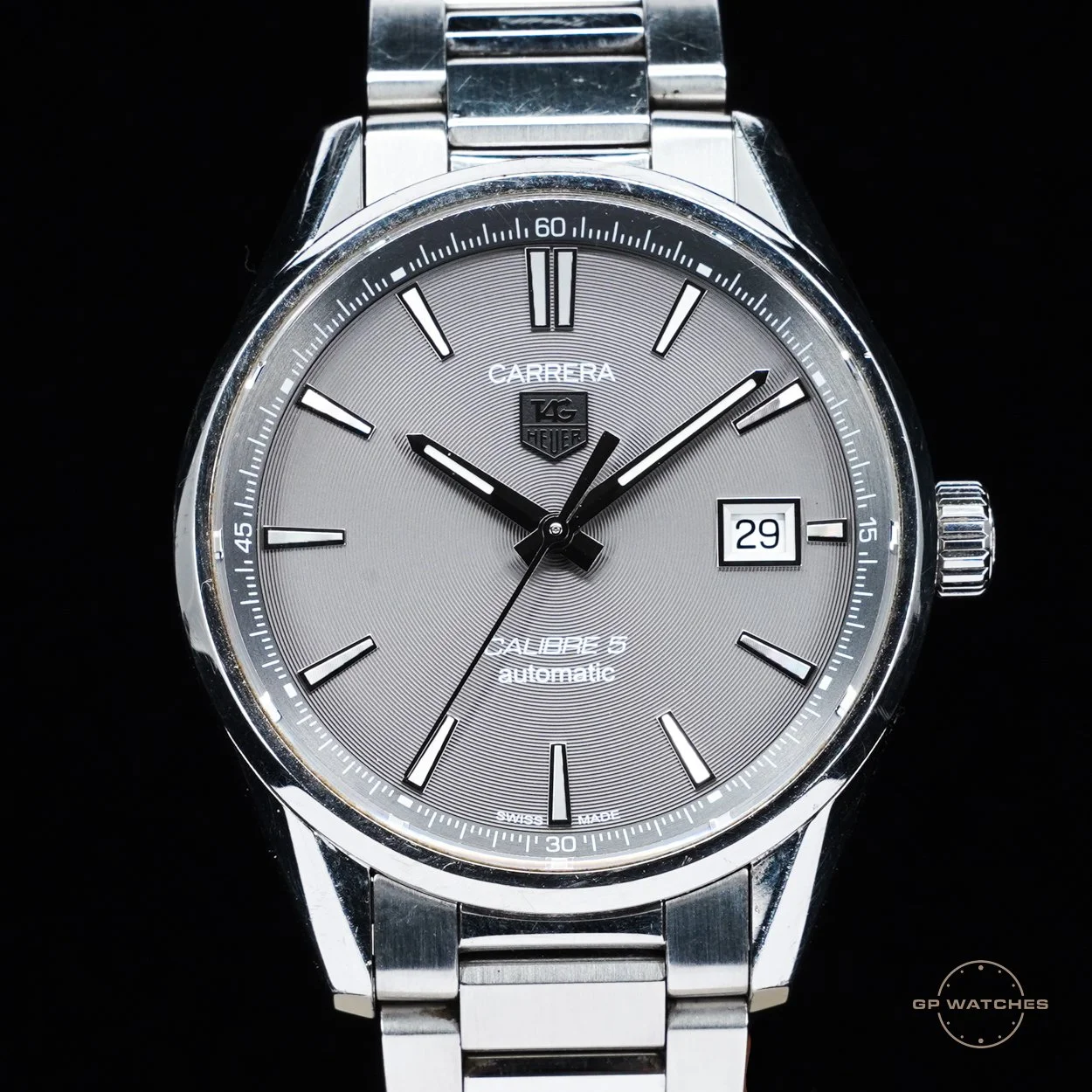 A close-up of a silver TAG Heuer Carrera Calibre 5 automatic wristwatch with a silver dial, black hands, date window at 3 o'clock, and a stainless steel bracelet.