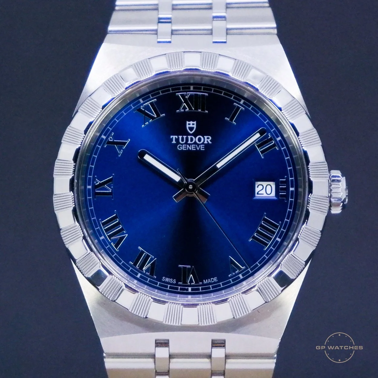 A close-up of a Tudor Geneve wristwatch with a blue dial, Roman numerals, a date display at 3 o'clock, and a stainless steel bracelet.