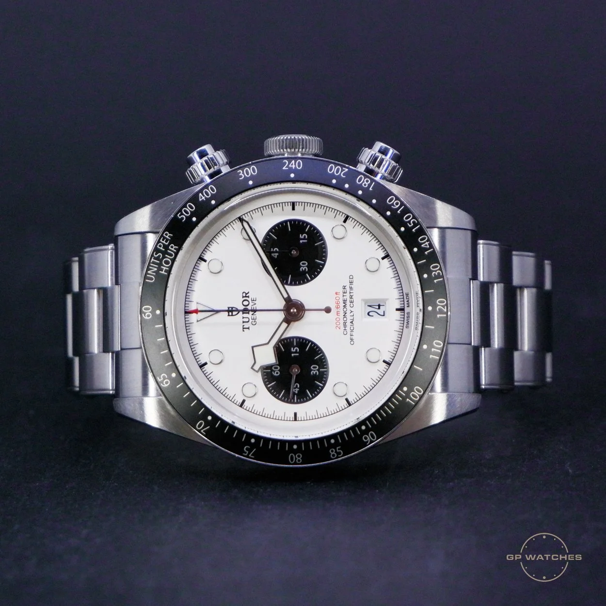 A Tudor chronograph watch with a white dial, black subdials, and a black bezel with tachymeter markings, silver bracelet, and date display at 3 o'clock.