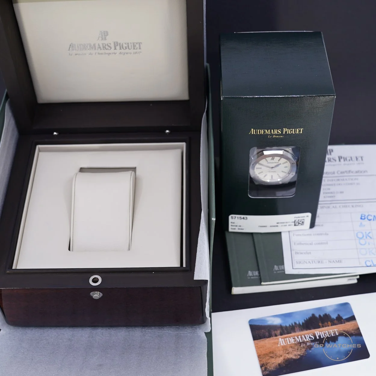 A luxury Audemars Piguet watch inside its open display box, with the watch in front of the original box and packaging, along with authentication paperwork and a promotional card.