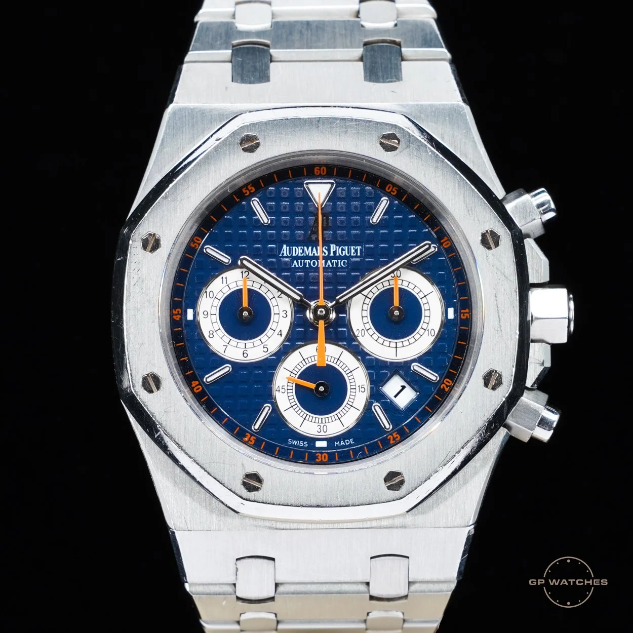 A luxury stainless steel Audemars Piguet chronograph watch with a blue textured dial, three sub-dials, a date window, and orange accents on a black background.