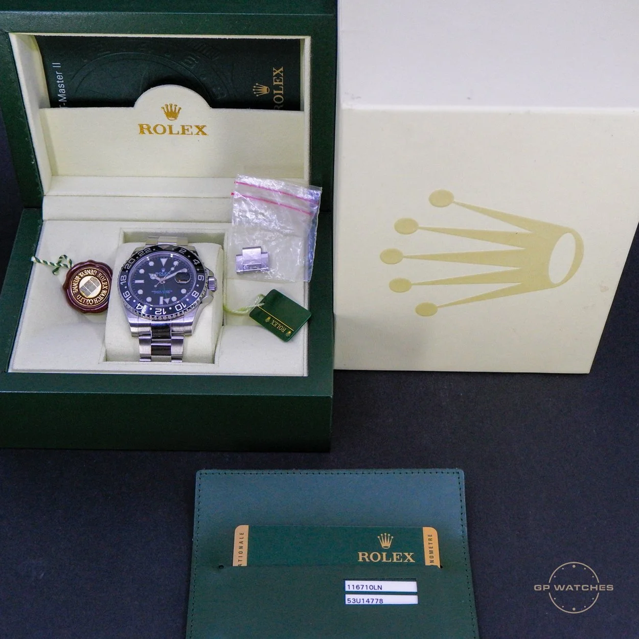 A Rolex watch inside a green and cream box, with a tag, extra links in a small plastic bag, and a Green Rolex tag. The outside of the box has a gold crown logo.