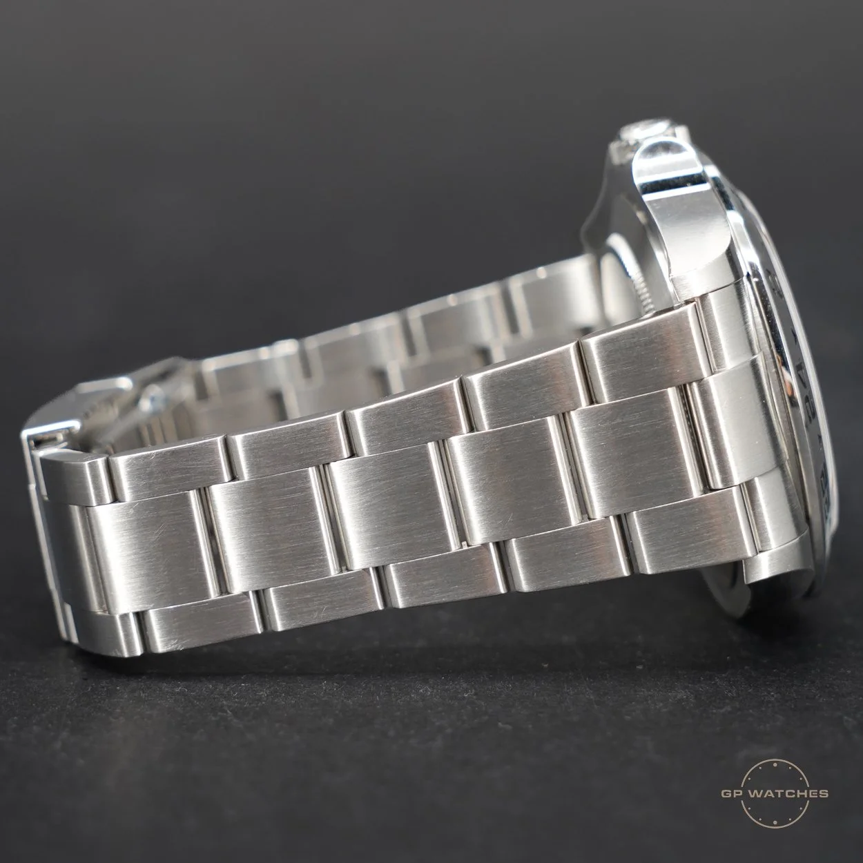 A stainless steel wristwatch with a brushed metal bracelet laying on a black surface. The watch face is partially visible, showing a silver bezel and some of the dial. A small logo for GP Watches is in the bottom right corner.