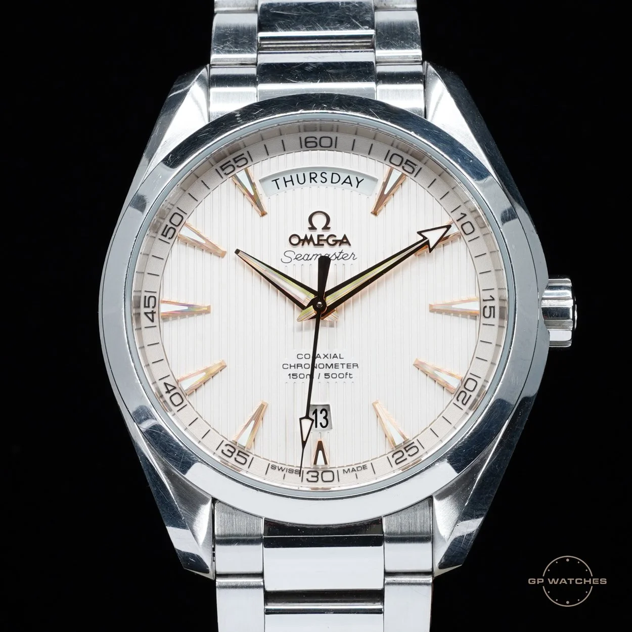 Close-up of an Omega Seamaster wristwatch with a white dial, gold hour markers, and a metal bracelet. The watch shows the day, date, and time, with the time indicating 10:10:30.