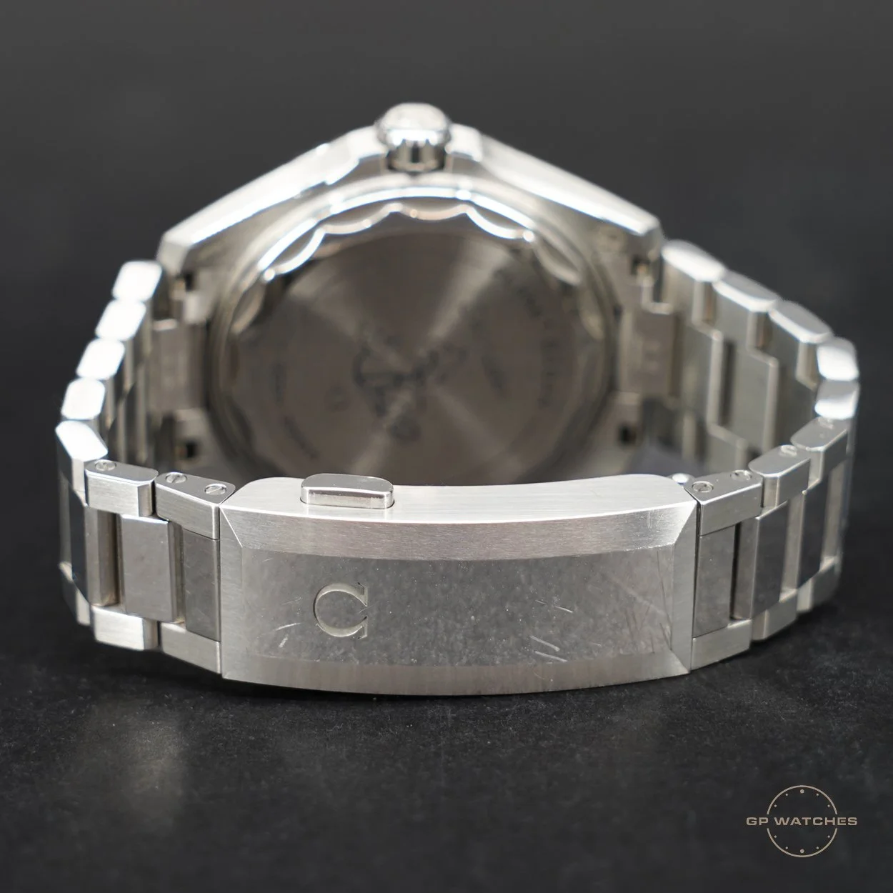 Close-up view of a silver wristwatch with a metal link bracelet, showing the back of the watch and the clasp engraved with a 'C'. The watch face is partially visible, and the background is black.