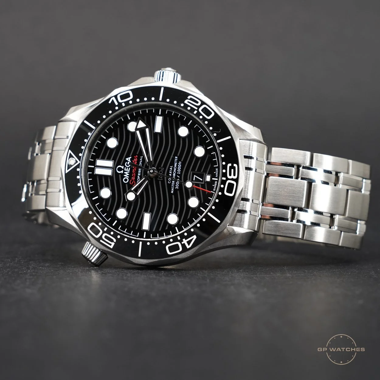 A close-up photograph of an Omega Seamaster wristwatch with a black dial, white luminous hour markers, and a stainless steel bracelet. The watch features a rotating bezel, thick hour and minute hands, and the date displayed at the 6 o'clock position. The background is dark gray with the logo 'GP Watches' in the bottom right corner.