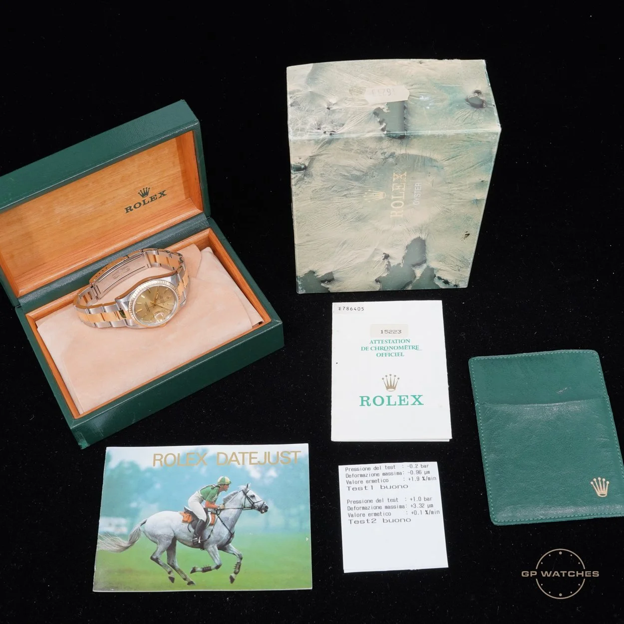 A Rolex Datejust watch with a gold dial and two-tone metal bracelet inside an open green and wooden box, a Rolex logo card, a Rolex book with a horse racing image, a green leather wallet with the Rolex crown logo, and a testing printout with pressure and deformation readings on a black surface.