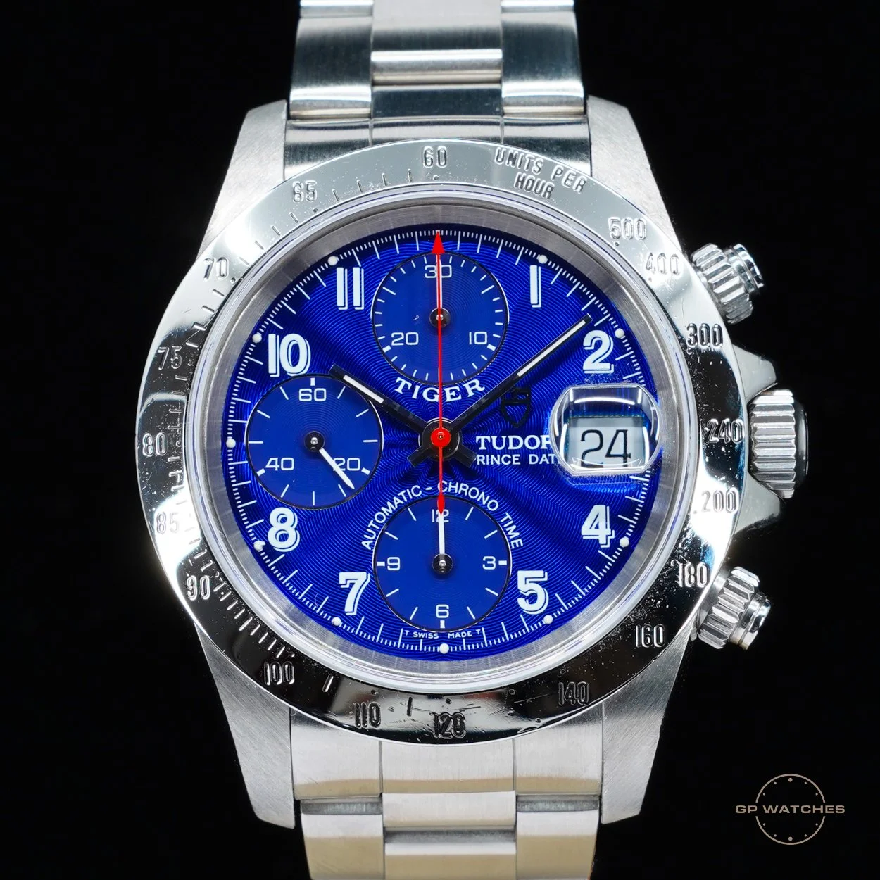 A close-up of a Tudor Prince Date chronograph wristwatch with a blue dial, silver bezel, and a date display at 3 o'clock, featuring multiple subdials and white and red hands.