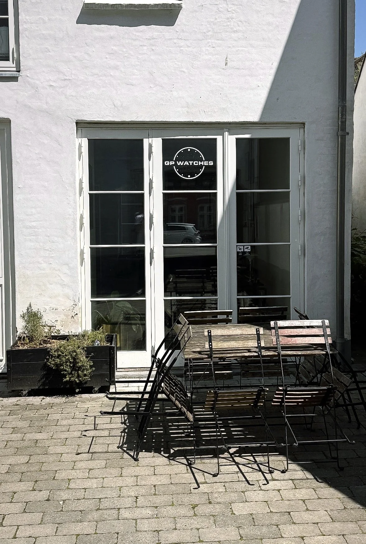 Outdoor view of a building with a door and window. Sign on the window reads 'GP WATCHES.' There are stacked wooden and metal chairs and a table in front, with a potted plant to the left on a brick patio.