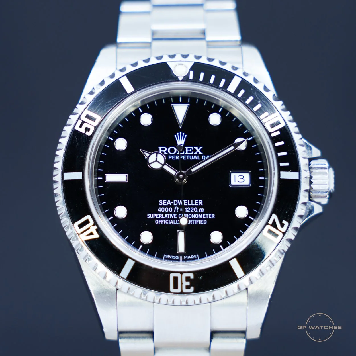 Close-up of a Rolex Sea-Dweller wristwatch with a black dial, stainless steel bracelet, and a rotating black and silver bezel, displaying the date at 3 o'clock, on a dark background.