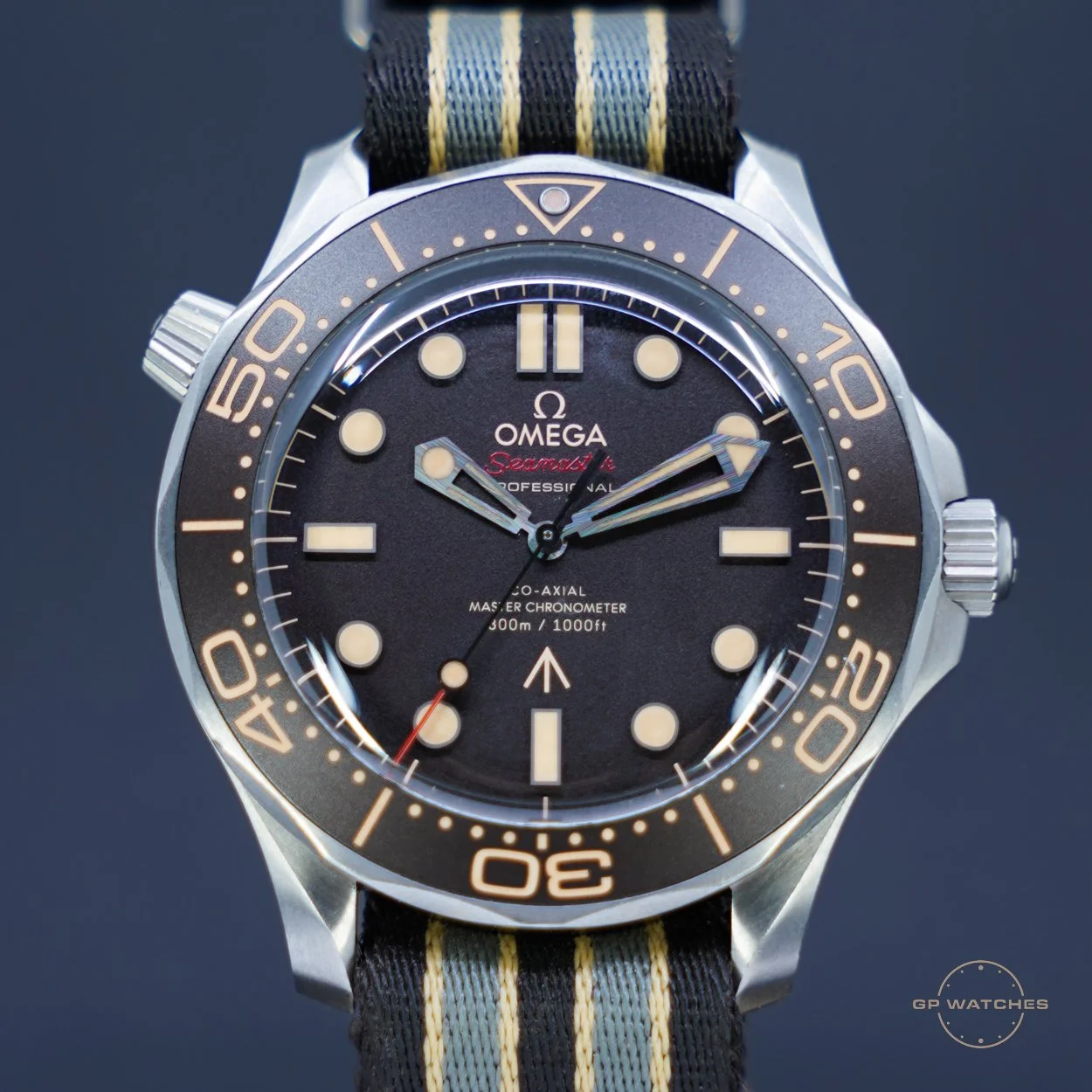 A close-up of an Omega Seamaster Professional dive watch with a black dial, luminous hour markers, and a rotating bezel with markers for diving. The watch has a fabric strap with navy, gray, and yellow stripes.