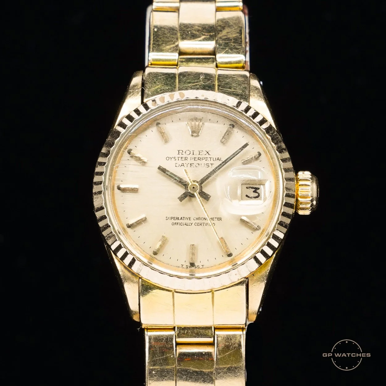 A gold Rolex Oyster Perpetual Datejust wristwatch with a beige dial, gold hour markers, and a date window at 3 o'clock with a magnifying lens, against a black background.