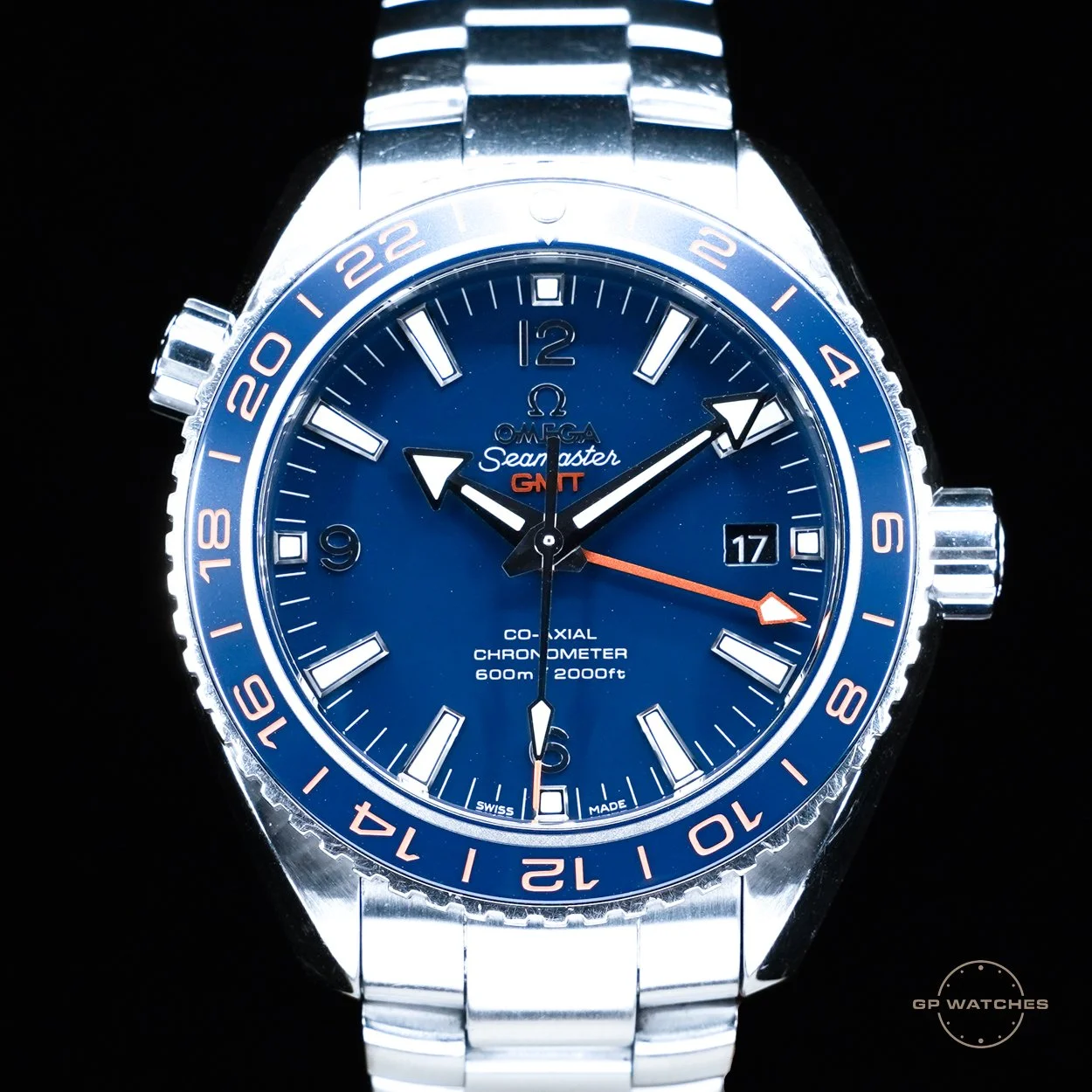A close-up of an Omega Seamaster GMT wristwatch with a blue dial, silver bezel, and a stainless steel bracelet, showing the date as the 17th.