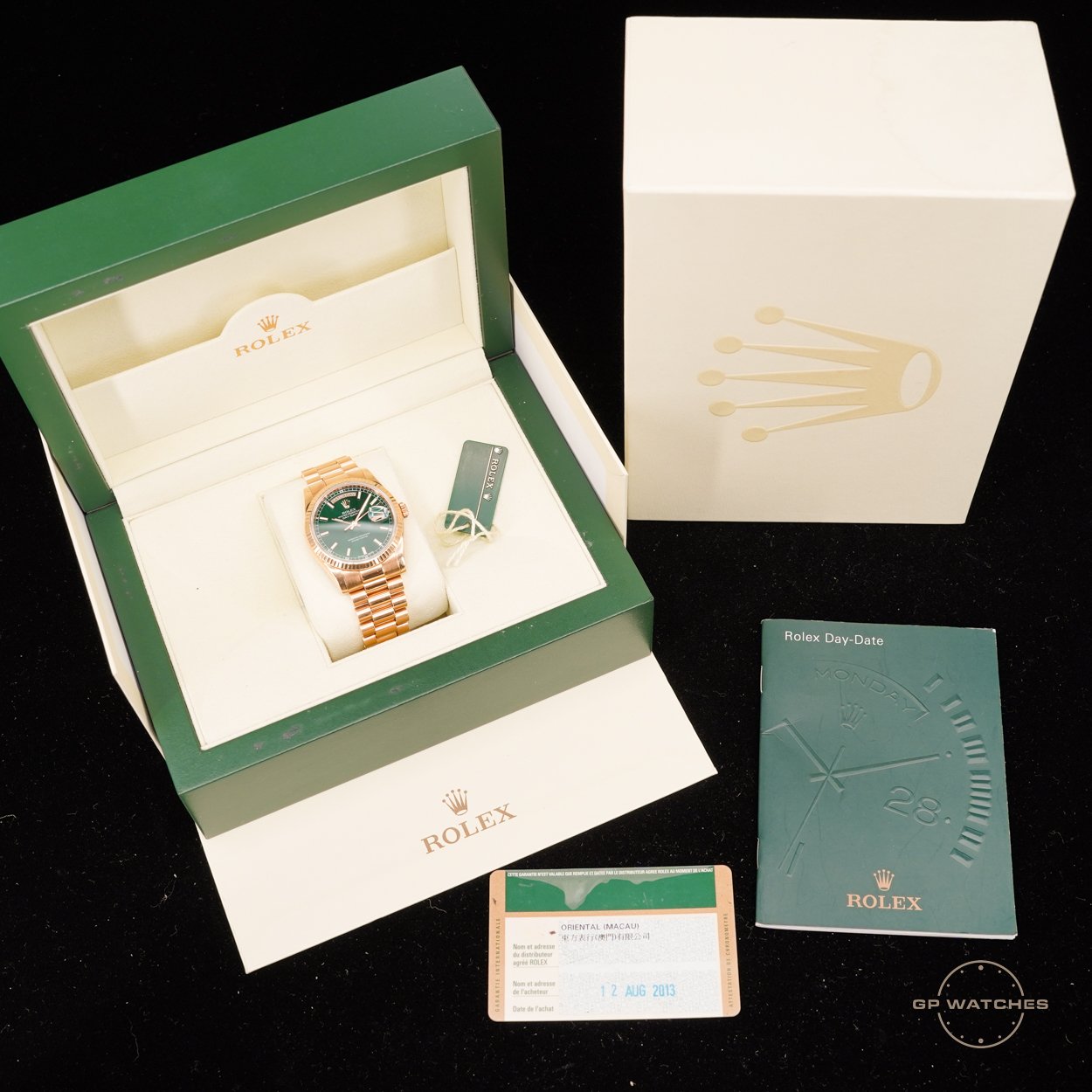 A Rolex Day-Date wristwatch with a green dial and gold bracelet inside its green and white box, accompanied by documentation, a tags, and a white outer box with a gold crown logo, all placed on a black surface.