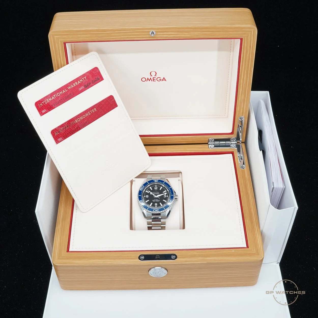 Omega wristwatch with a black dial and blue bezel inside a wooden display box, with warranty and certification cards.