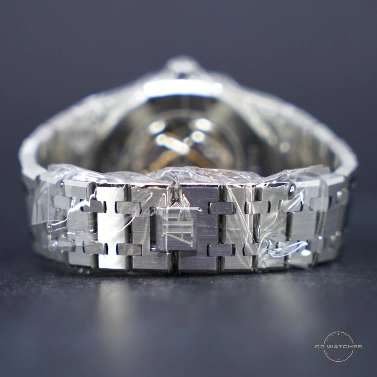 Close-up of a silver wristwatch with a metal band, partially covered in clear plastic protective film, against a dark background, with the GP Watches logo in the bottom right corner.