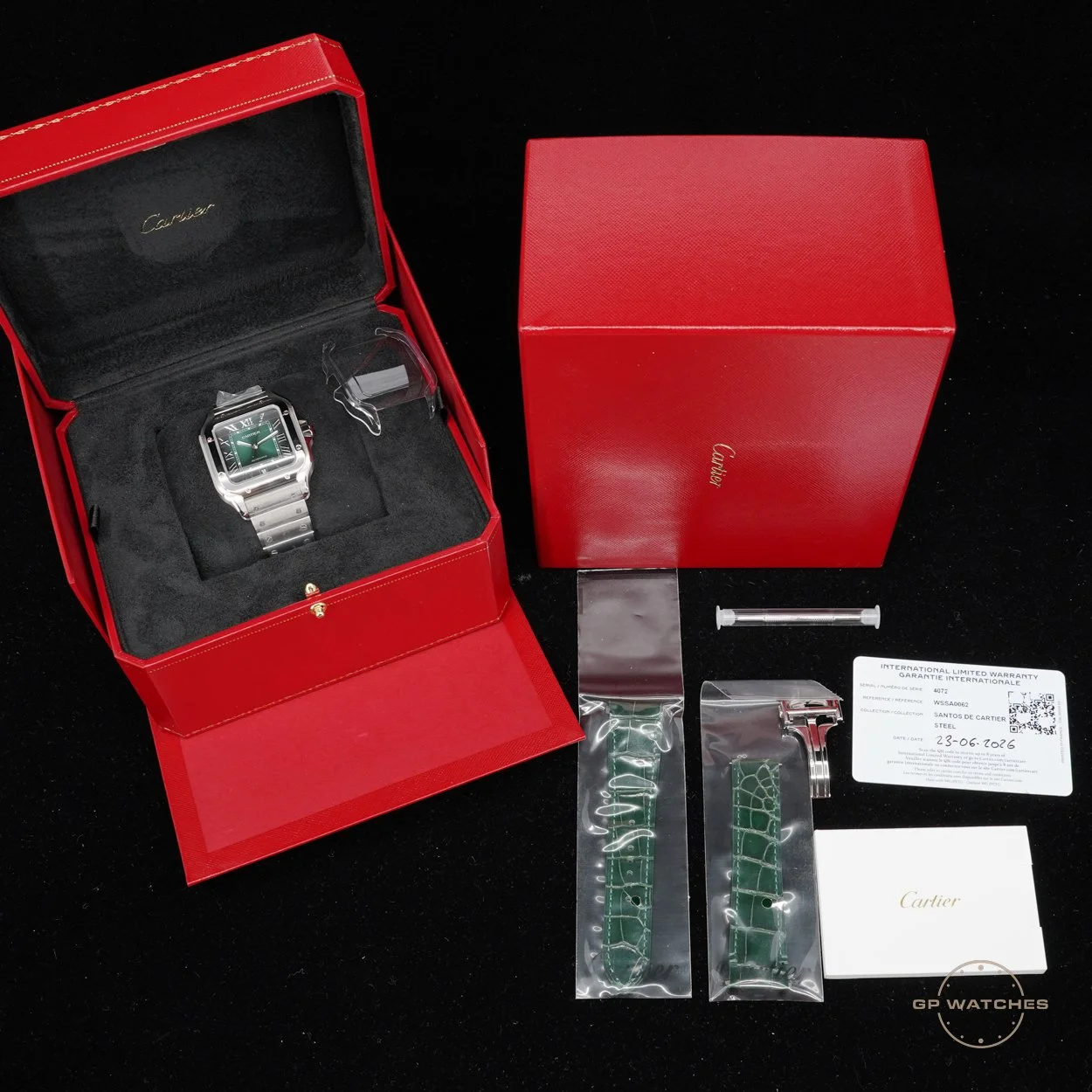 A silver Cartier wristwatch with a green dial in its red Cartier box, accompanied by extra green watch straps, a small syringe-shaped tool, a warranty card, and a white booklet, all on a black surface.