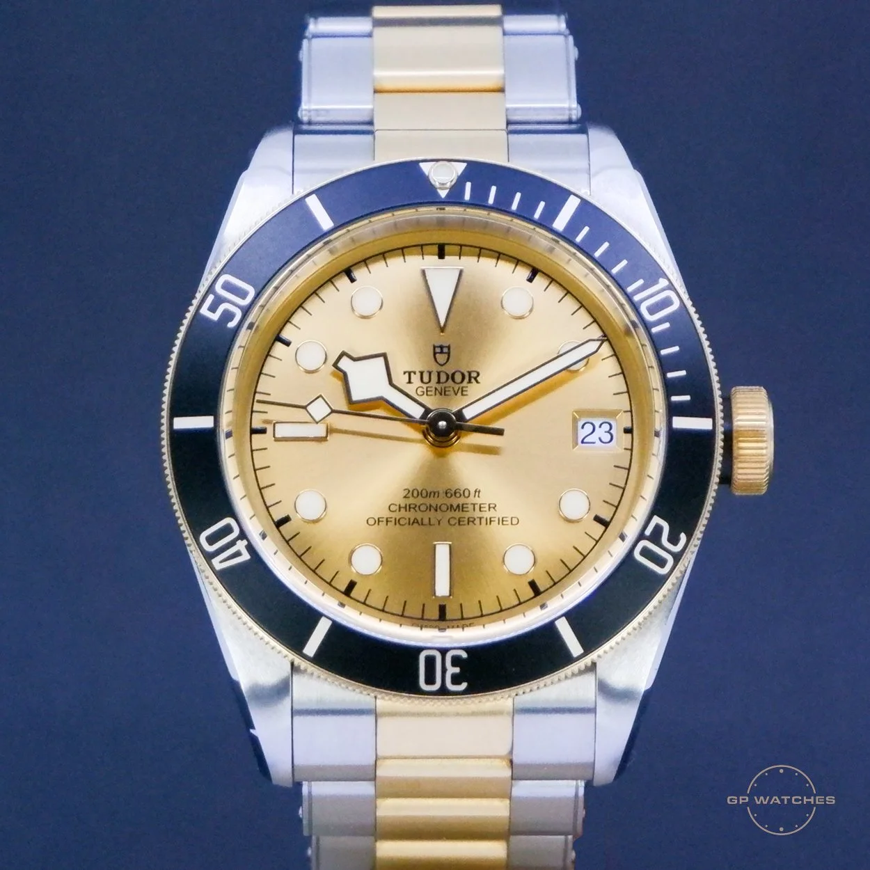 Two-tone Tudor wristwatch with gold and silver bracelet, gold dial, black and gold bezel, date window at 3 o'clock, and three hands, set against a dark background.