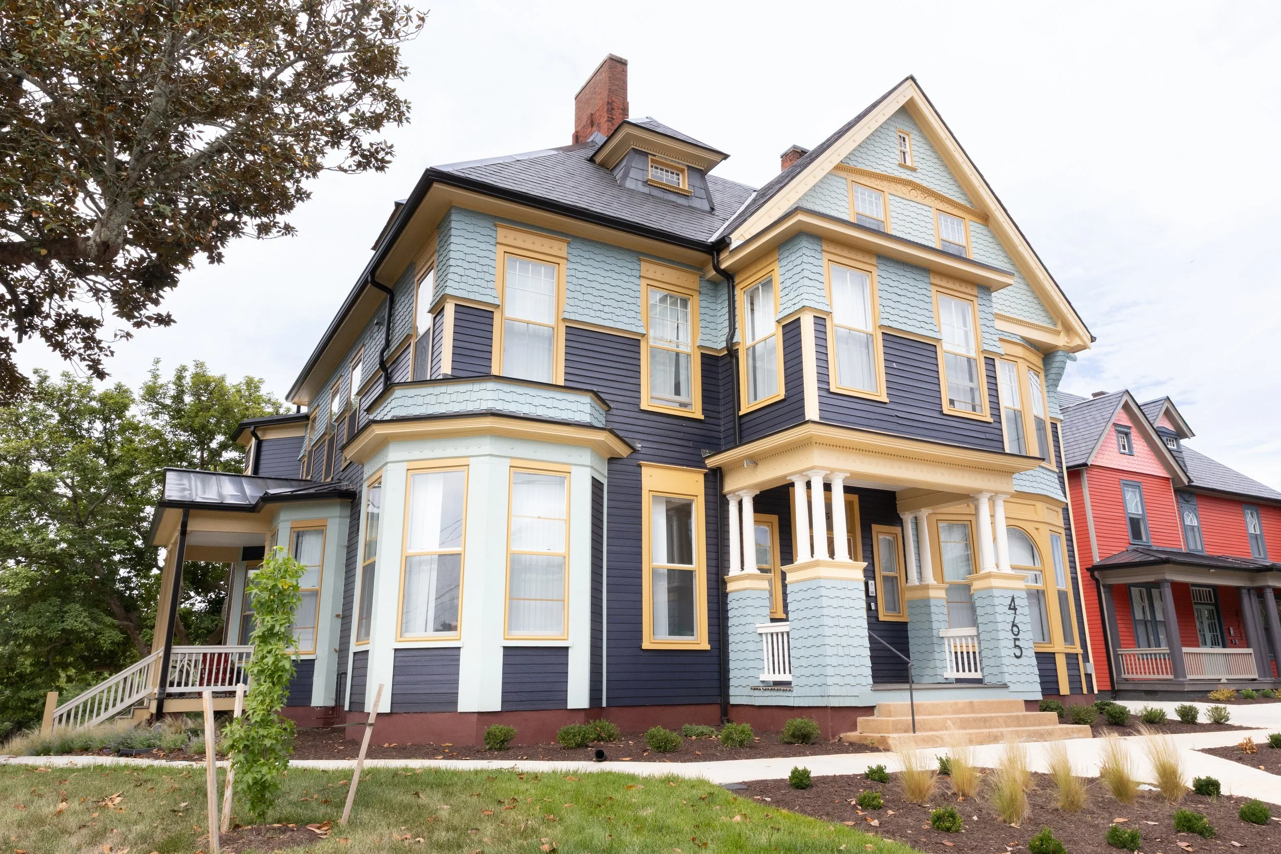 A large, colorful Victorian-style house with blue and yellow paint, multiple windows, and a porch, set in a landscaped yard with small bushes and trees.