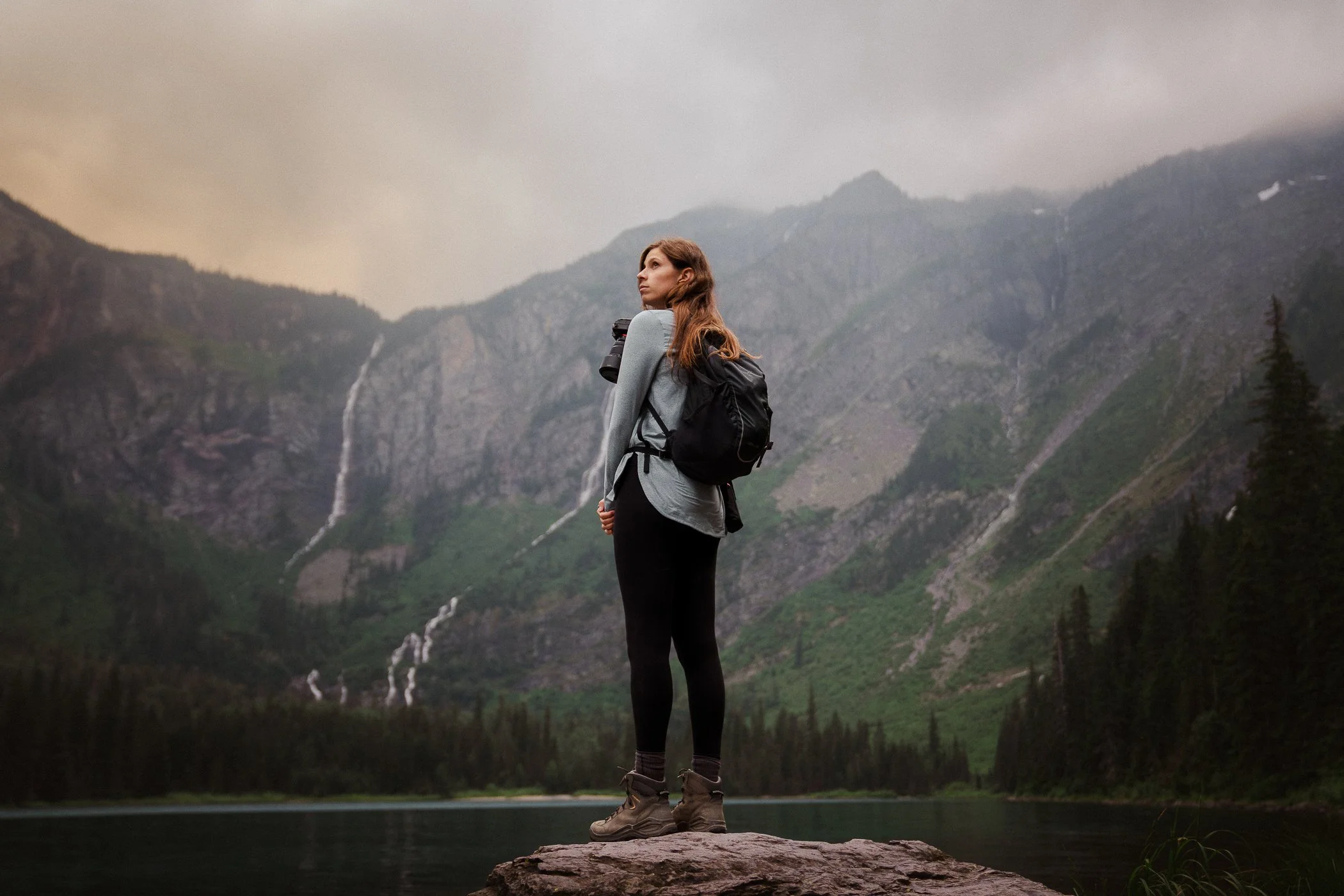 Hiker woman standing on a rock in Glacier National Park with a small pack, camera, and bear spray