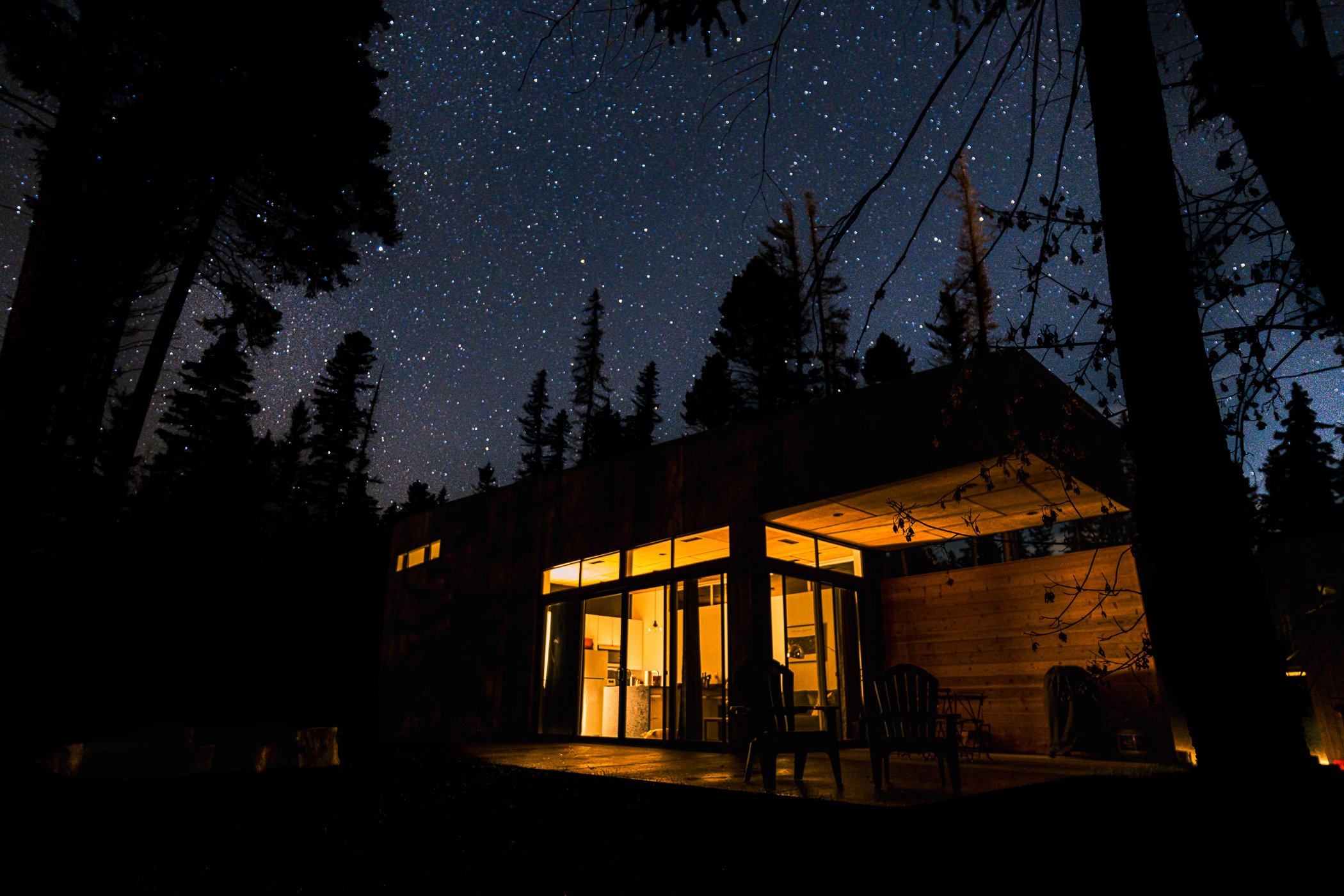 Nighttime view of a house with large glass windows and warm lighting, seen through trees with star-filled sky in the background. Hospitality sample of staying at Stoner Creek Cabins overnight. Astrophoto sample.