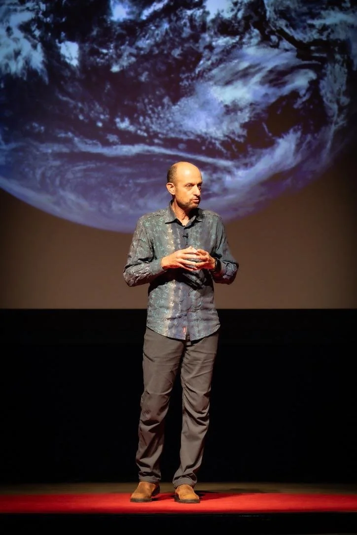 A man standing on stage with a large Earth image behind him, appearing to give a presentation or speech.