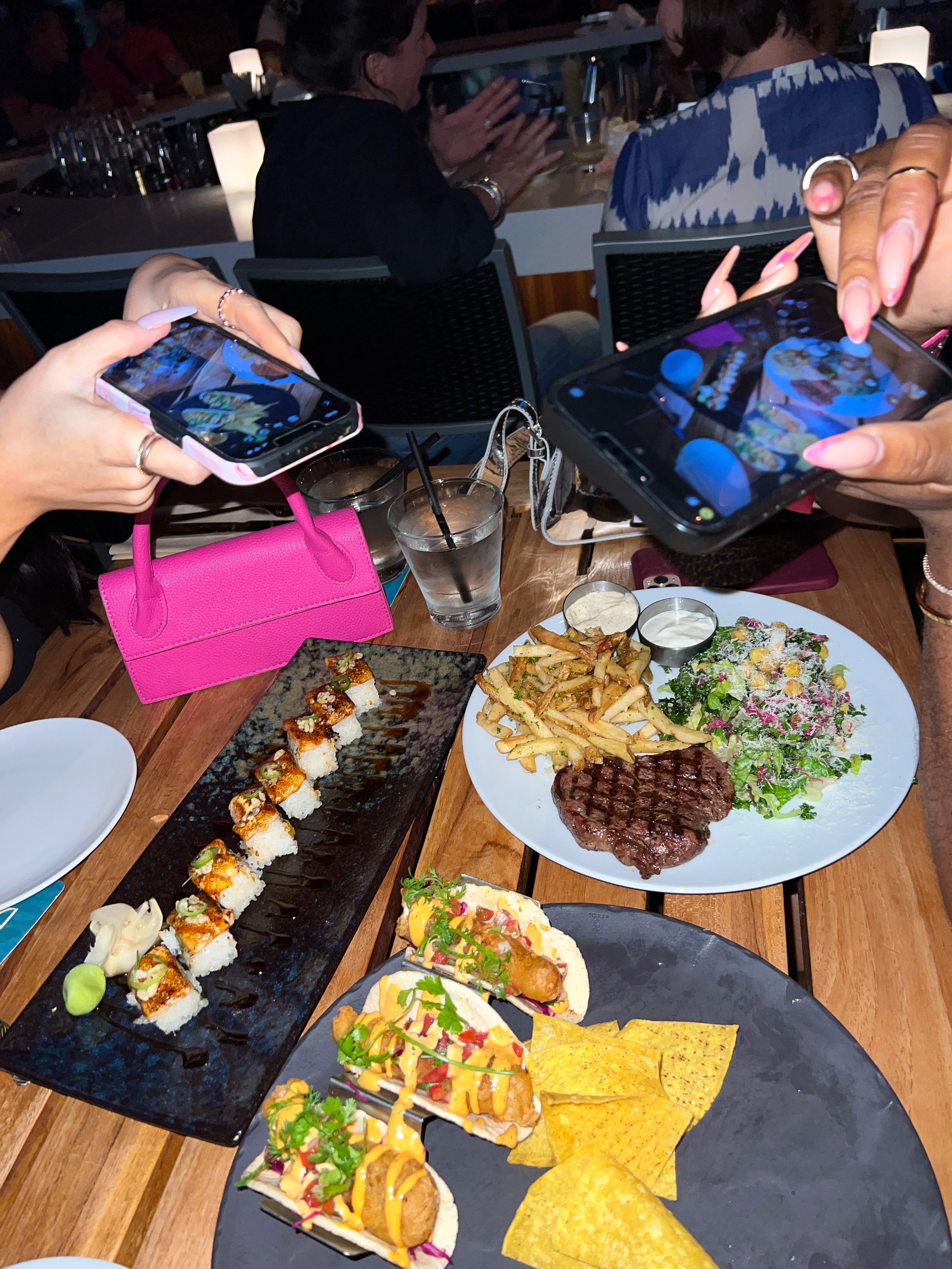 A table with various dishes including sushi rolls, steak with fries and salad, fish tacos, and tortilla chips, with people taking photos of the food using smartphones. A small pink purse is placed on the table.