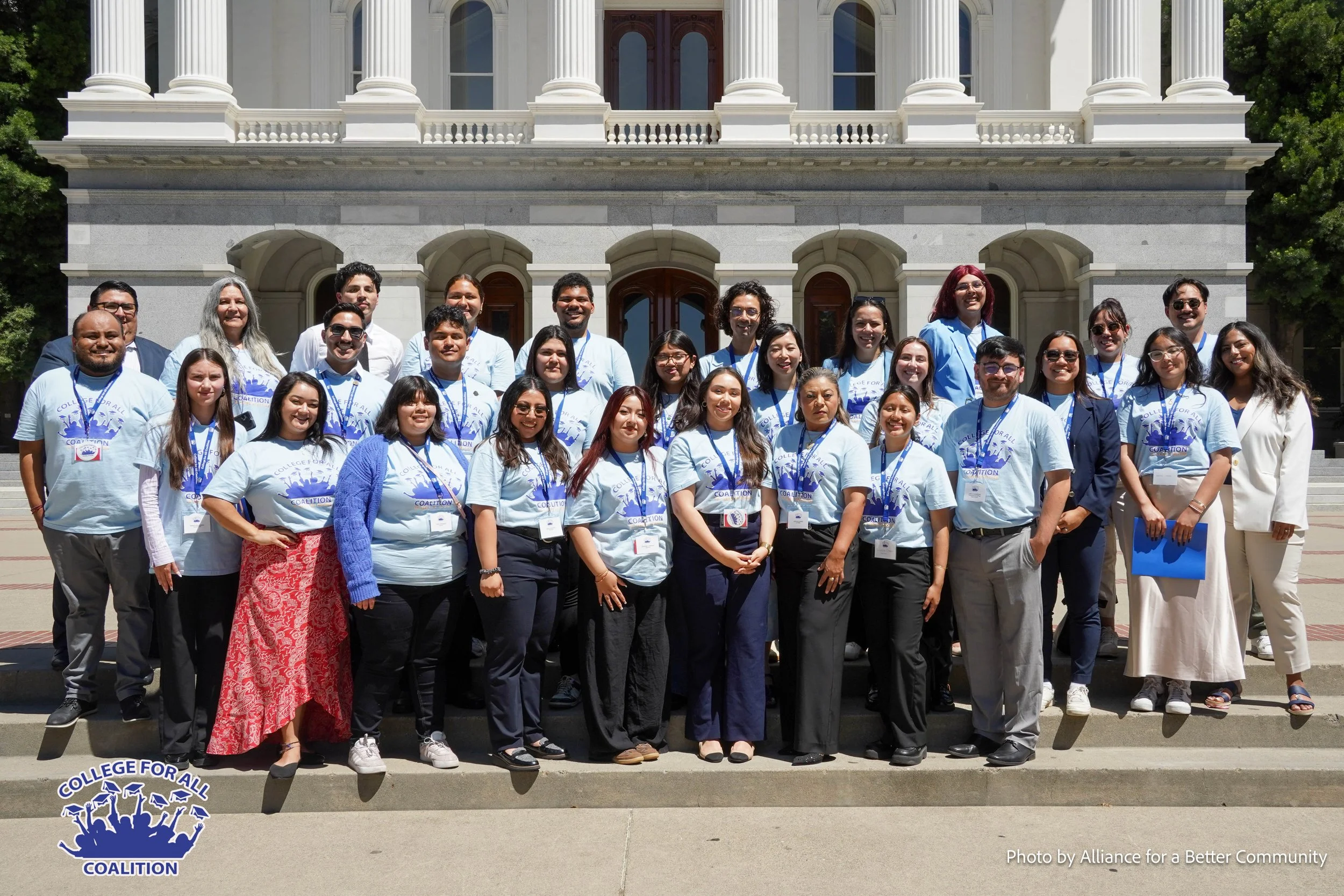 Group of diverse young people and adults standing on steps in front of a historic building, wearing light blue t-shirts with a logo that reads 'College for All Coalition.' The scene appears to be a group photo at a community event or rally.
