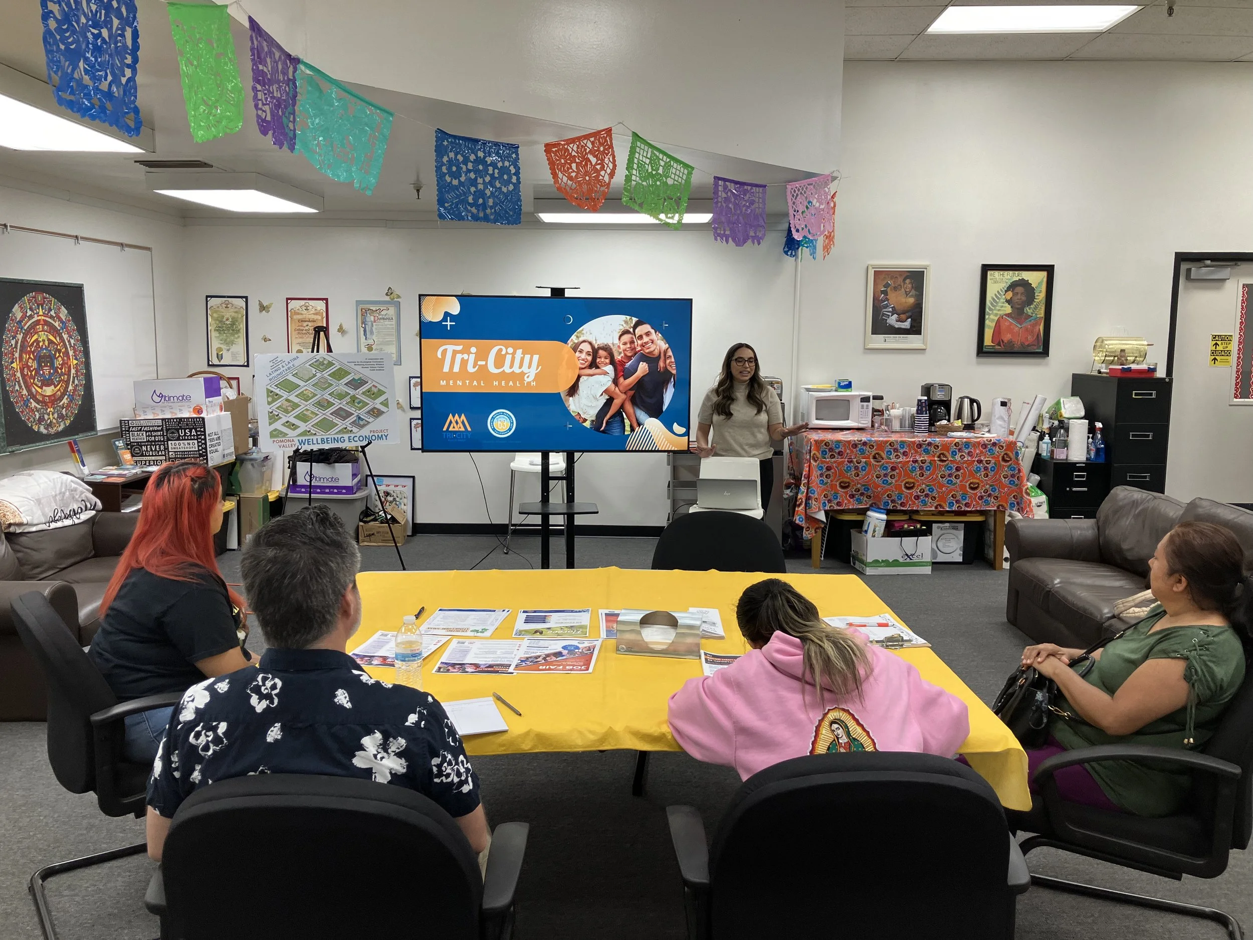 A group of four people attending a presentation in a conference room decorated with colorful papel picado banners. One woman is standing near a large screen displaying a slide about Tri-City Mental Health, and three attendees are seated facing her, listening and taking notes.