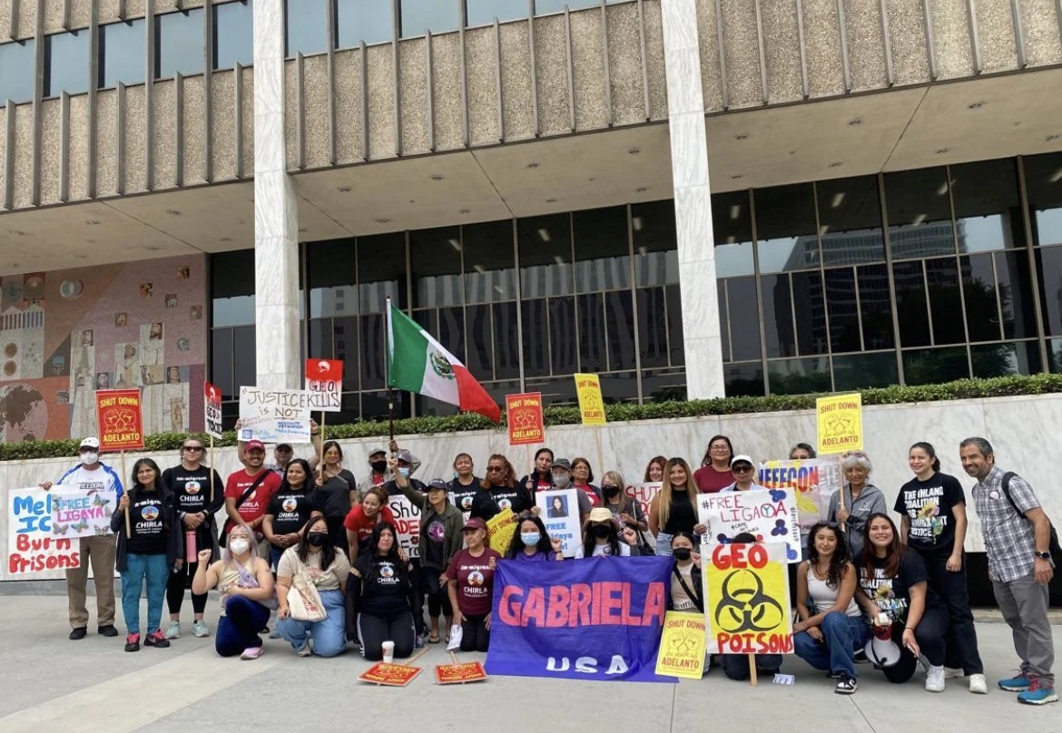 Group of people protesting outside a government building, holding signs with slogans like "Shut down Adelanto," "Justice for Tulis," "Free Ligaya," and "GABRIELA USA." Some signs have messages such as "Mel IC burn prisons" and "Geo poisons." A Mexican flag is visible among the protesters.