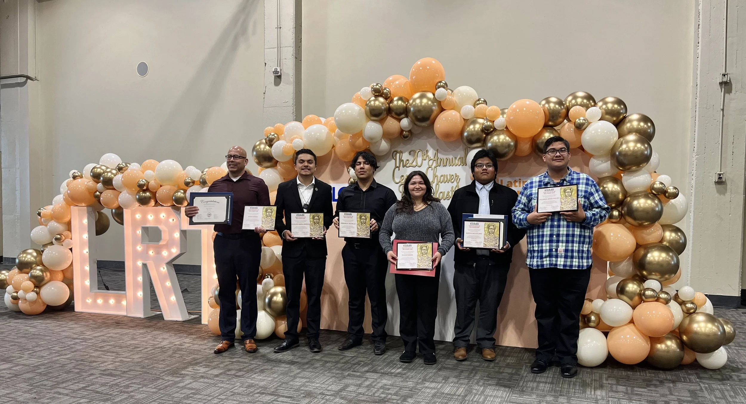 Six people standing in front of a balloon arch, holding certificates at a graduation ceremony.