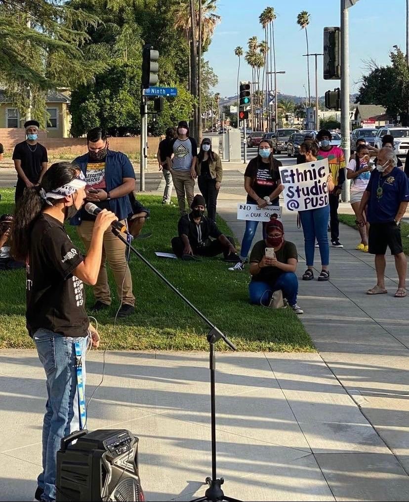 A young person with a headband is speaking into a microphone at a protest or rally, with a group of people standing and sitting behind them. Some of the attendees are holding signs, one of which reads "Ethnic Studies @ PUSD." The protest is outdoors, near a street with palm trees and traffic lights. Most people are wearing masks.