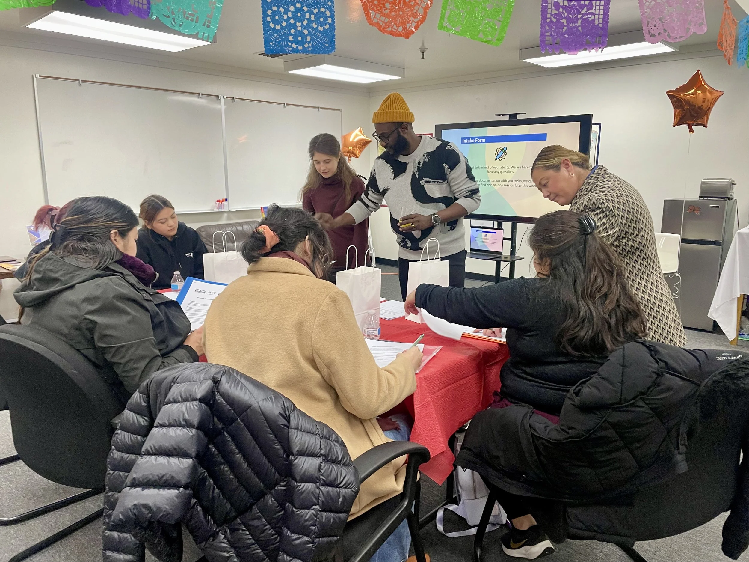 Group of people sitting around a table with gift bags, participating in a workshop or seminar, with a man and woman standing at the front near a large monitor.