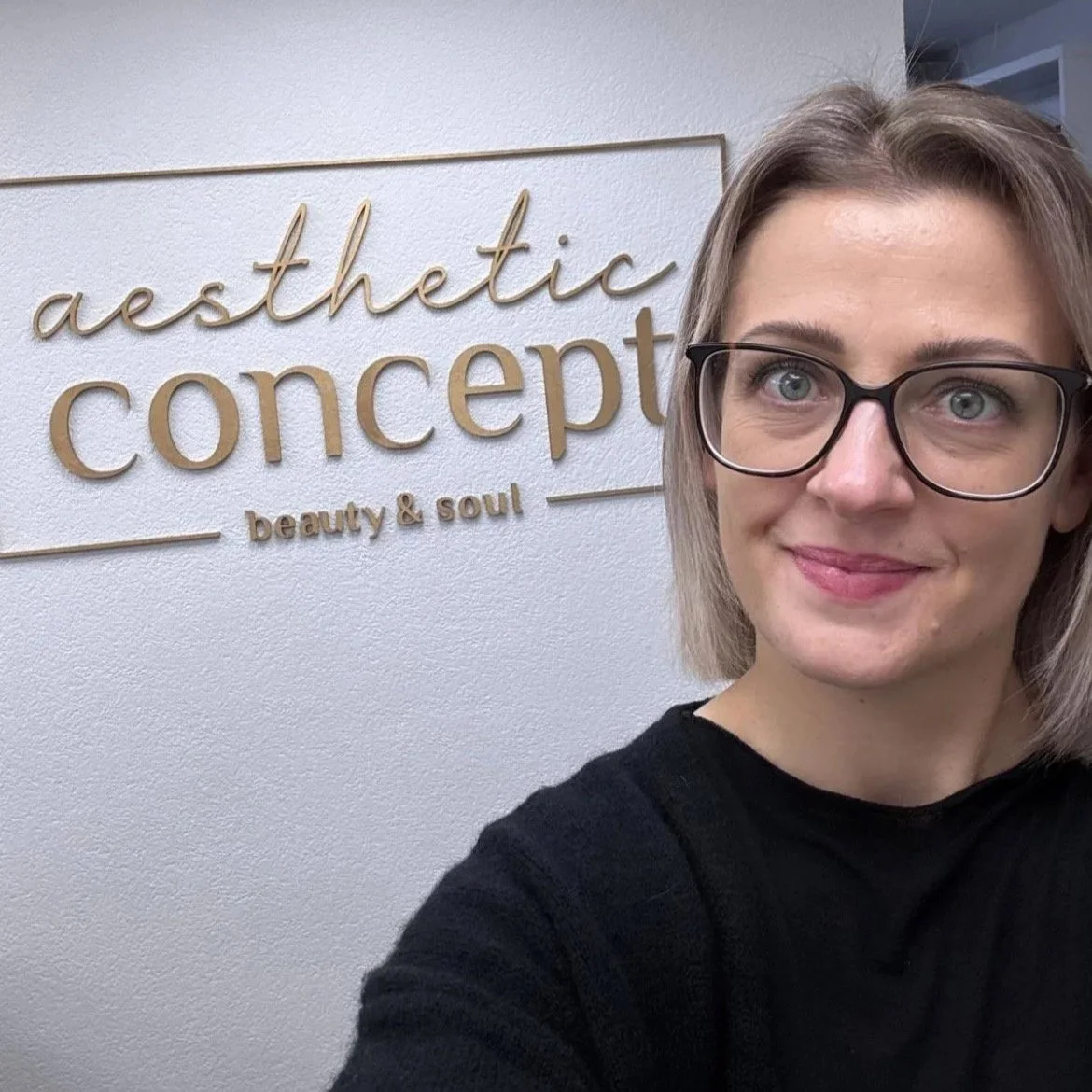 Ein lächelnder Frau mit Brille vor einem Wandlogo, das 'aesthetic concept beauty & soul' schreibt.