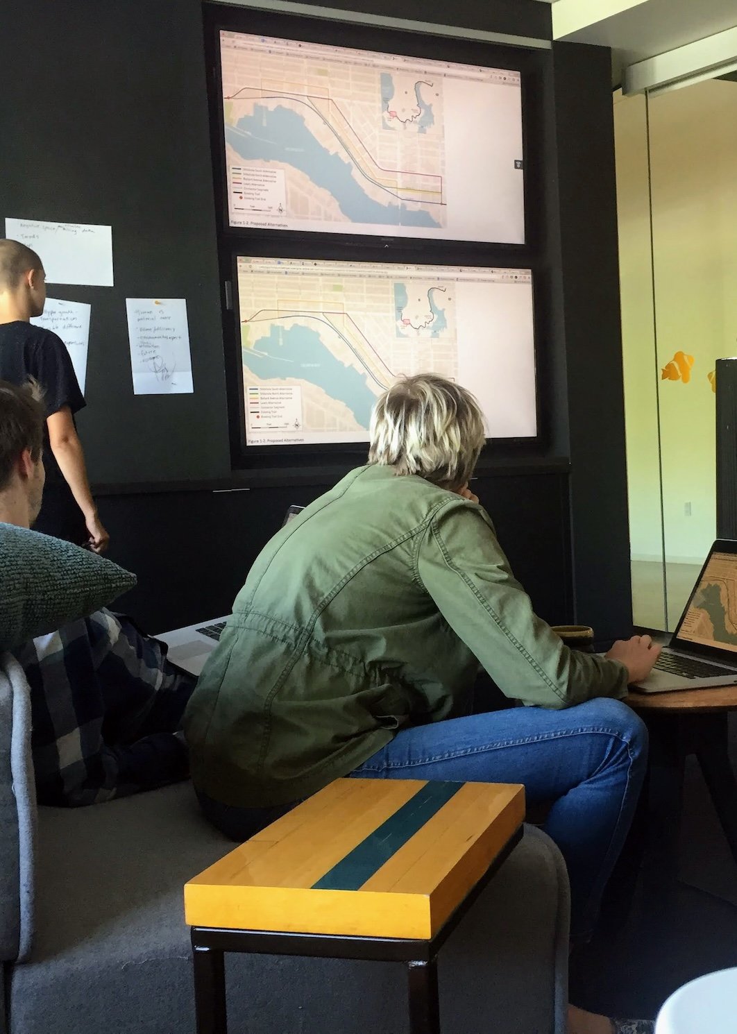 Three people in a conference room looking at wall-mounted displays showing maps