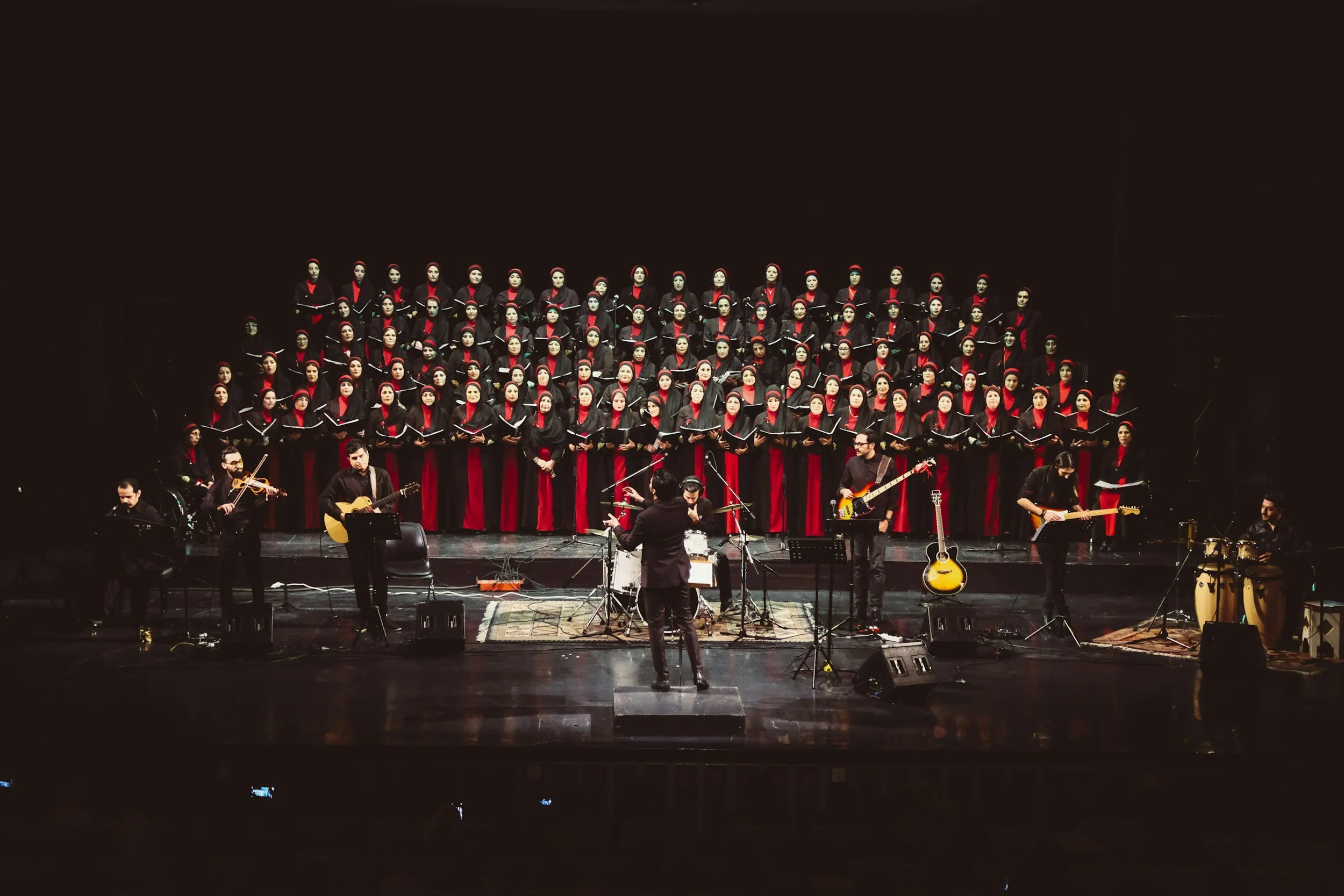 A large choir dressed in black and red robes performing on stage, accompanied by musicians playing guitars and percussion instruments in front of them.