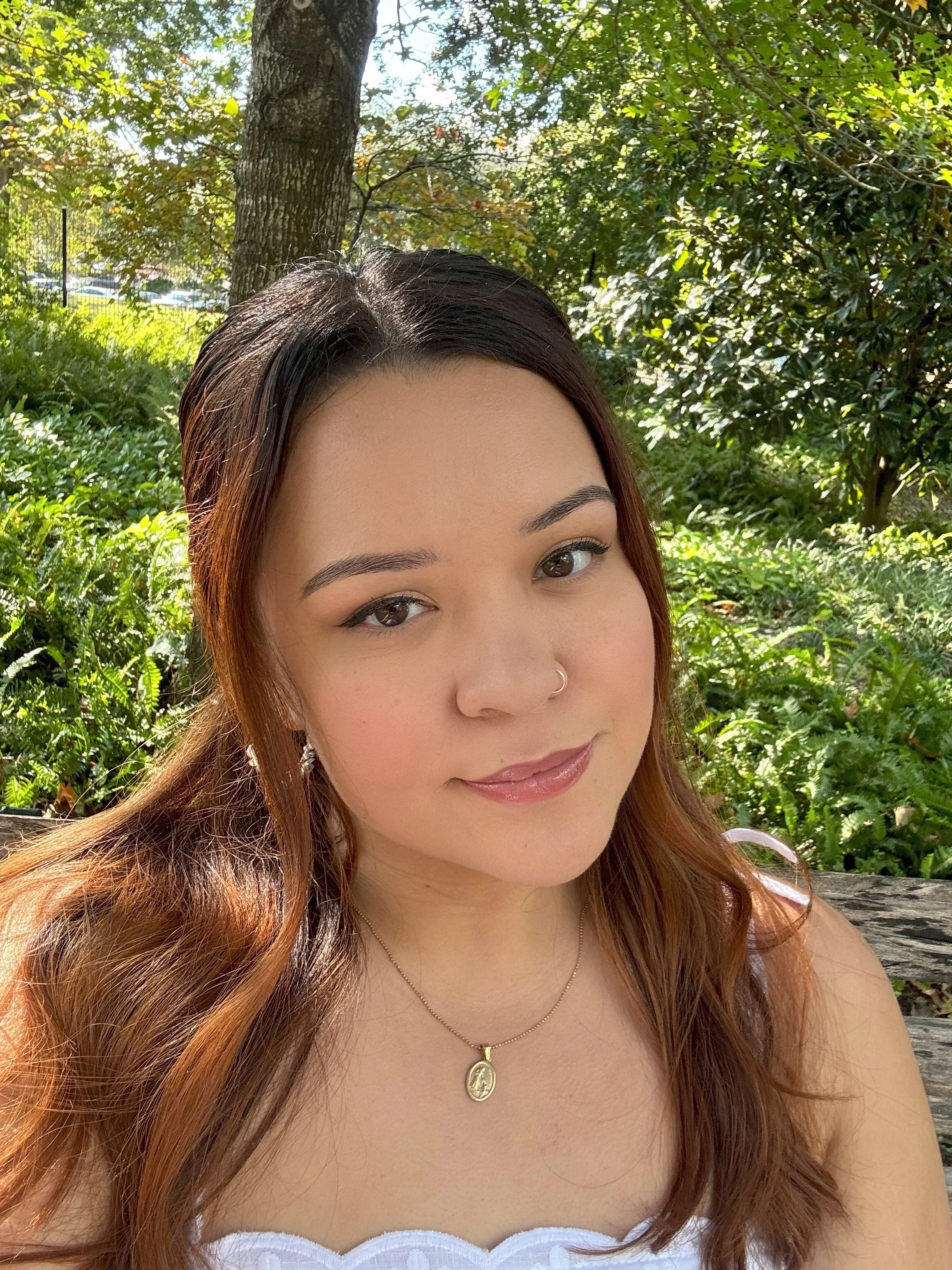 A young woman with long, wavy brown hair and light skin taking a selfie outdoors. She is wearing a white top, a gold necklace with a pendant, and a nose ring. She has makeup on and is smiling slightly. The background features green trees, bushes, and sunlight filtering through the leaves.