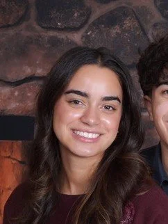 Close-up of a smiling woman with long dark hair, wearing a dark top, against a stone wall background.