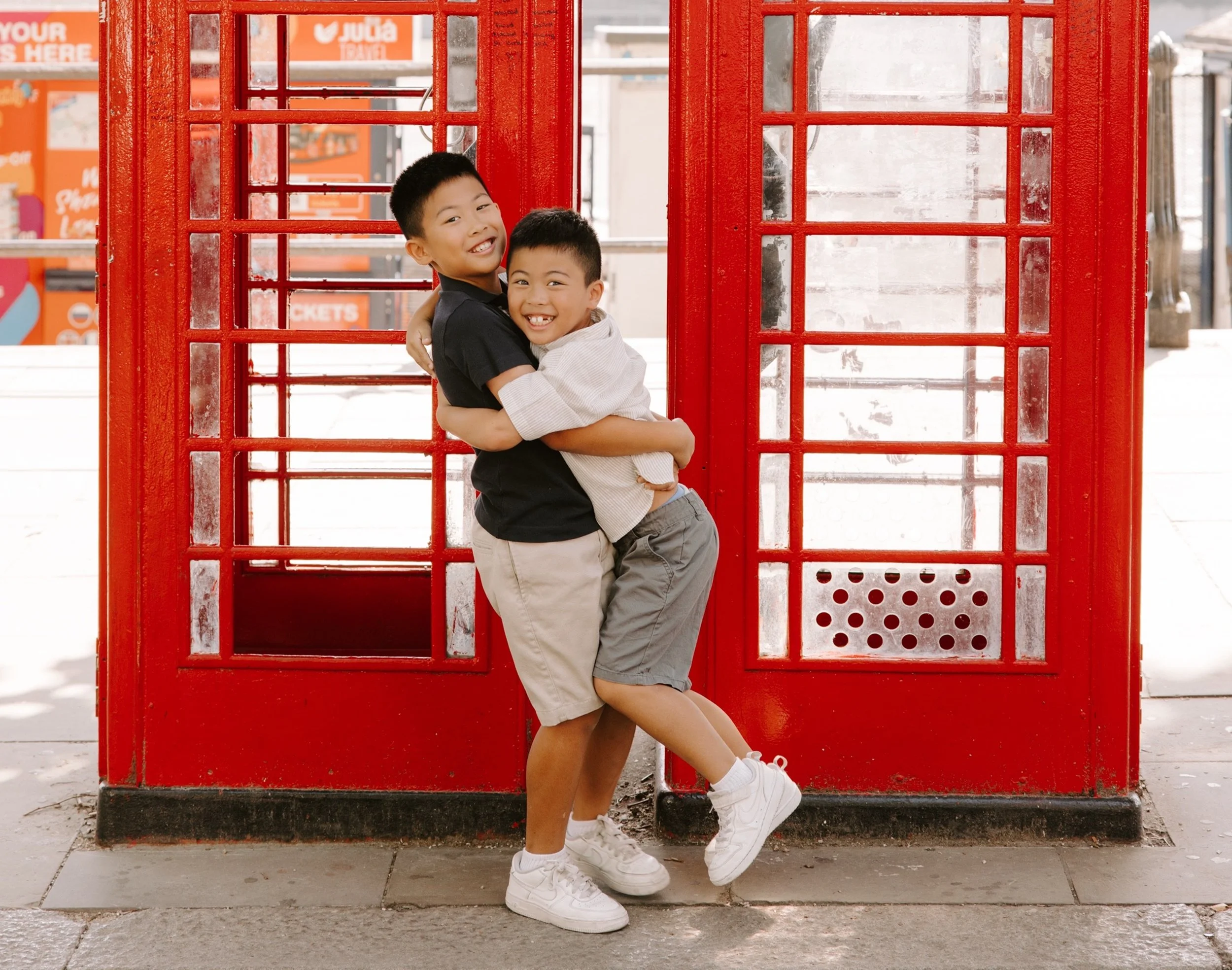 Two boys hugging in front of a red telephone booth on a sidewalk, smiling happily.