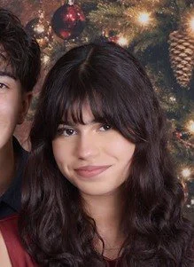 A young woman with dark wavy hair and bangs smiling in front of a decorated Christmas tree.